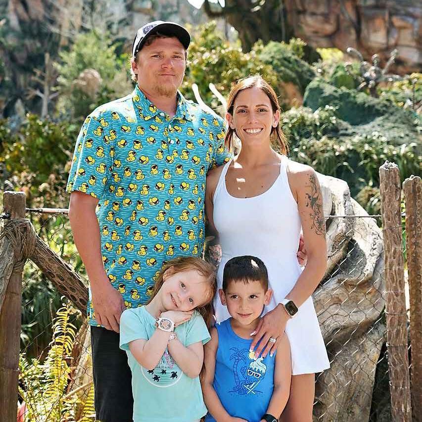 A smiling family of four poses together outdoors near a wooden fence and lush greenery, with two parents and two young children.
