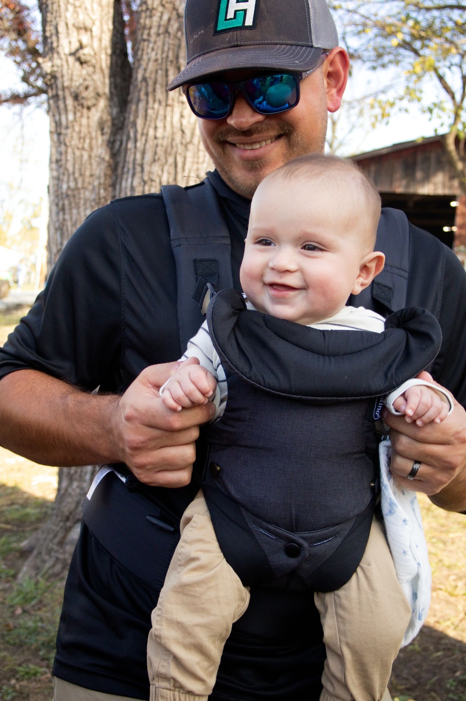 Smiling man in sunglasses and a baseball cap holds a happy baby facing outward in a black baby carrier outdoors near trees.