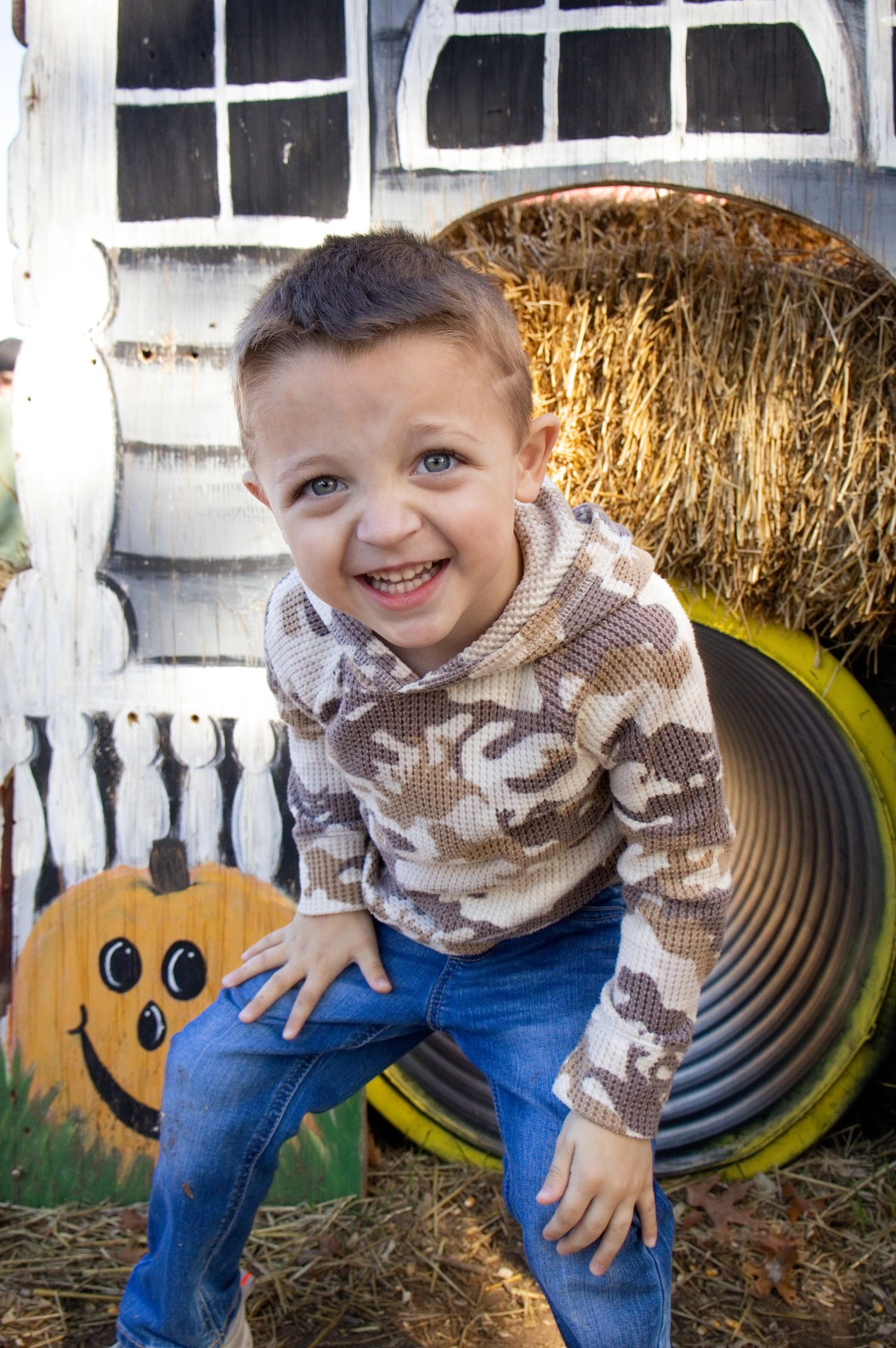 Smiling young boy in a camo hooded shirt and jeans sits by a hay bale and tunnel near a painted pumpkin at a fall pumpkin patch.