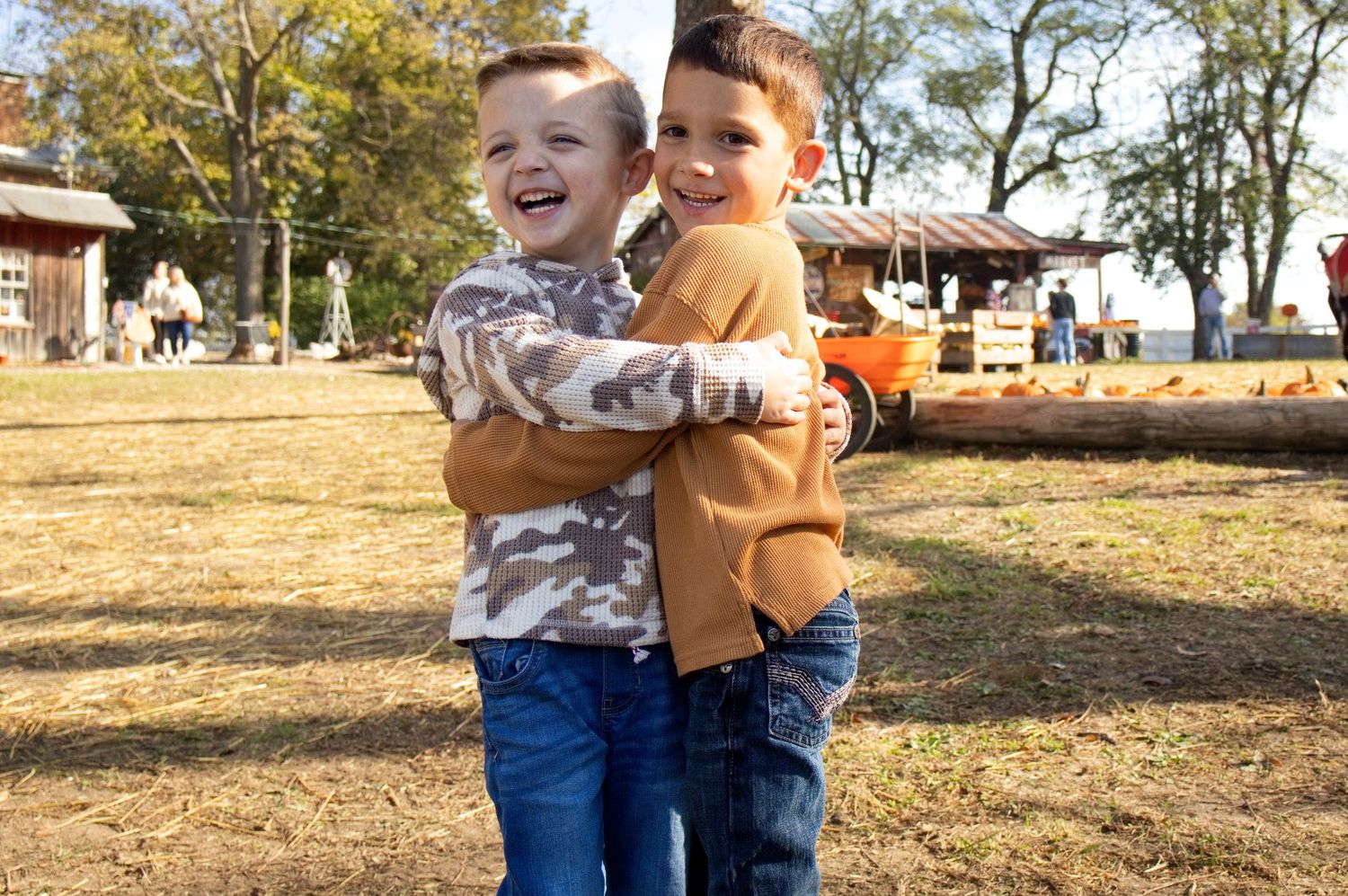 Two smiling young boys in jeans hug each other outdoors on a grassy field at a pumpkin patch with a rustic barn and trees behind them.