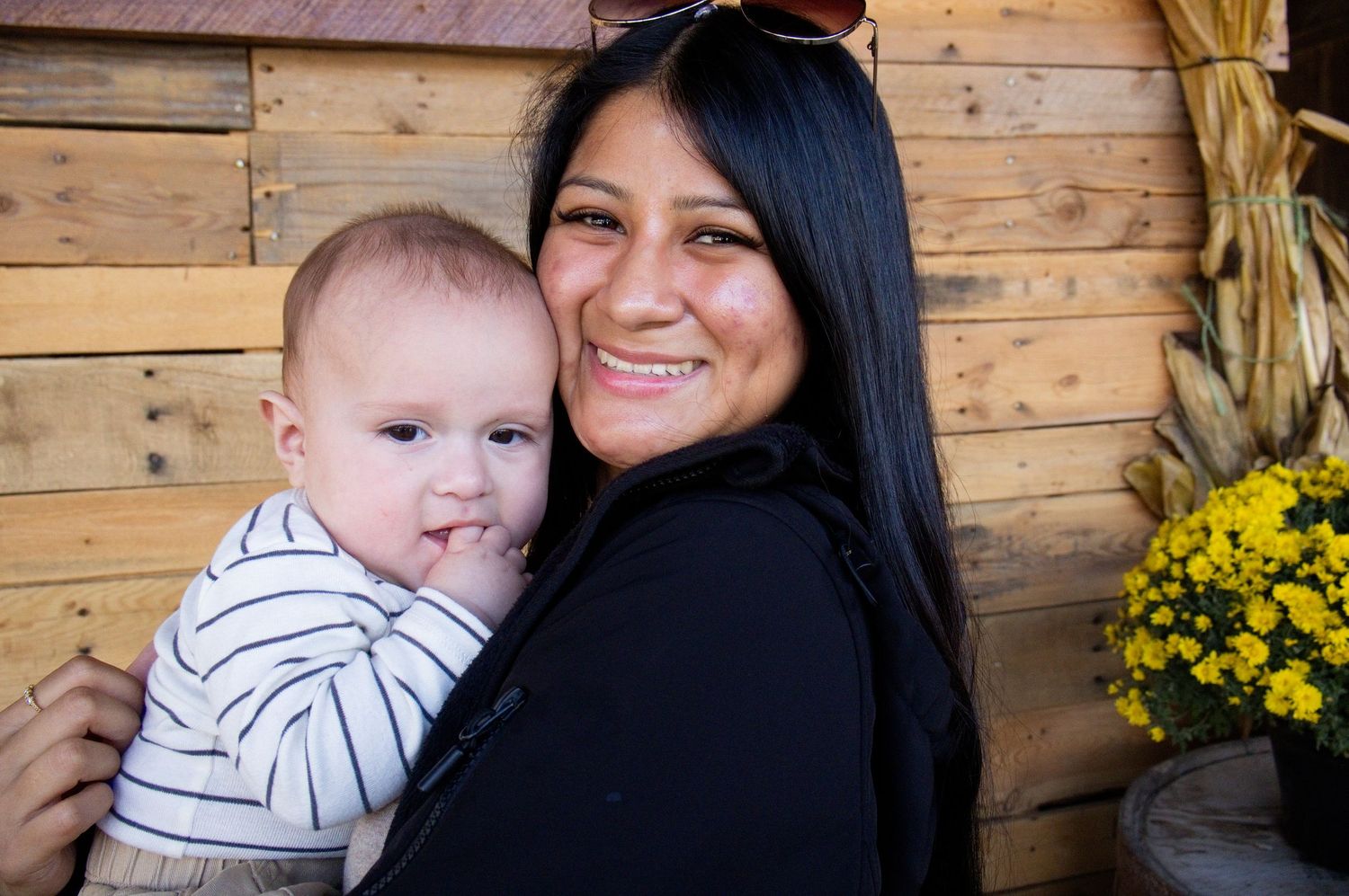 Smiling woman with long dark hair holds a baby in a striped shirt beside a wooden wall and yellow chrysanthemums with corn stalks.