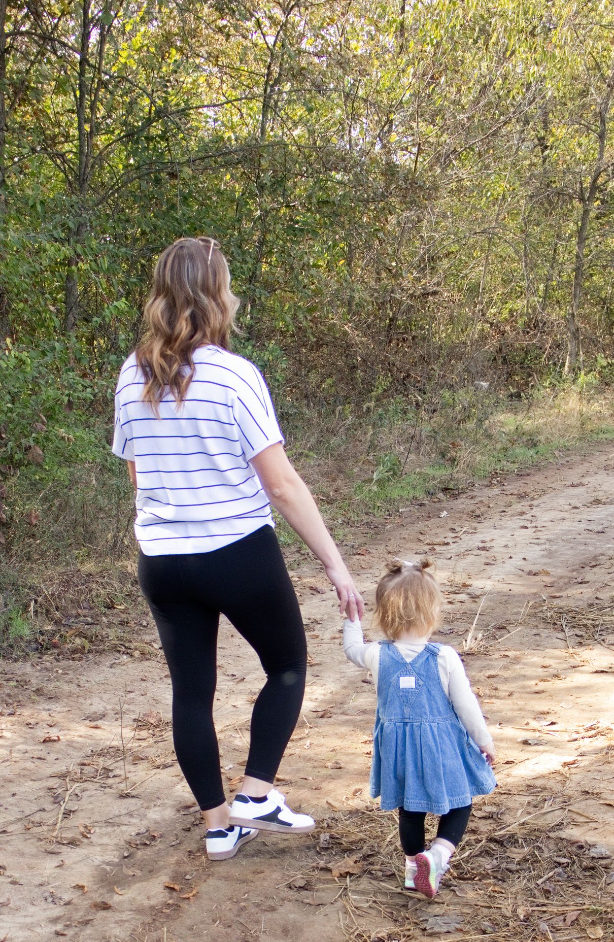Woman in a striped tee and black leggings holds hands with a toddler in a denim dress while walking down a dirt path through autumn woods.