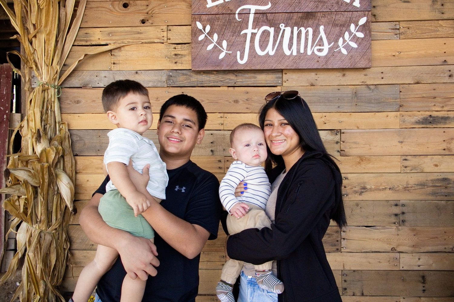 Smiling man and woman hold two young children in front of a rustic wooden wall with a Farms sign and dried corn stalks.