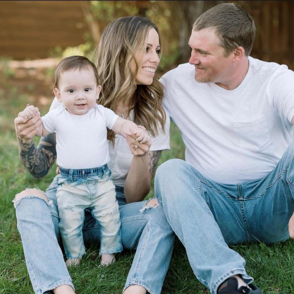 A smiling woman and man in white shirts and jeans sit on green grass holding the hands of a happy baby between them outdoors.