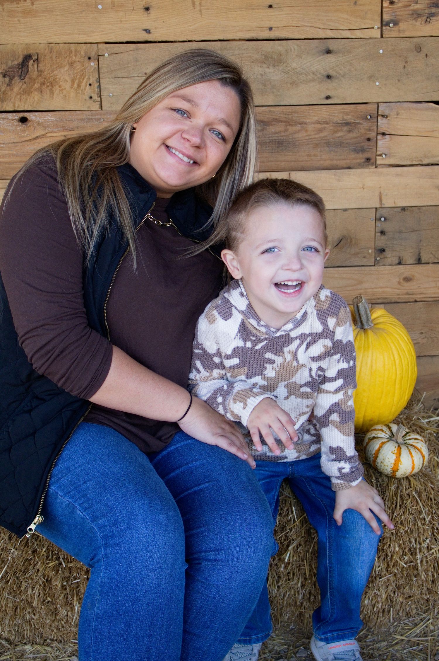 Smiling woman in a black vest sits on a hay bale with a laughing toddler in a camo hoodie beside pumpkins and rustic wood planks.