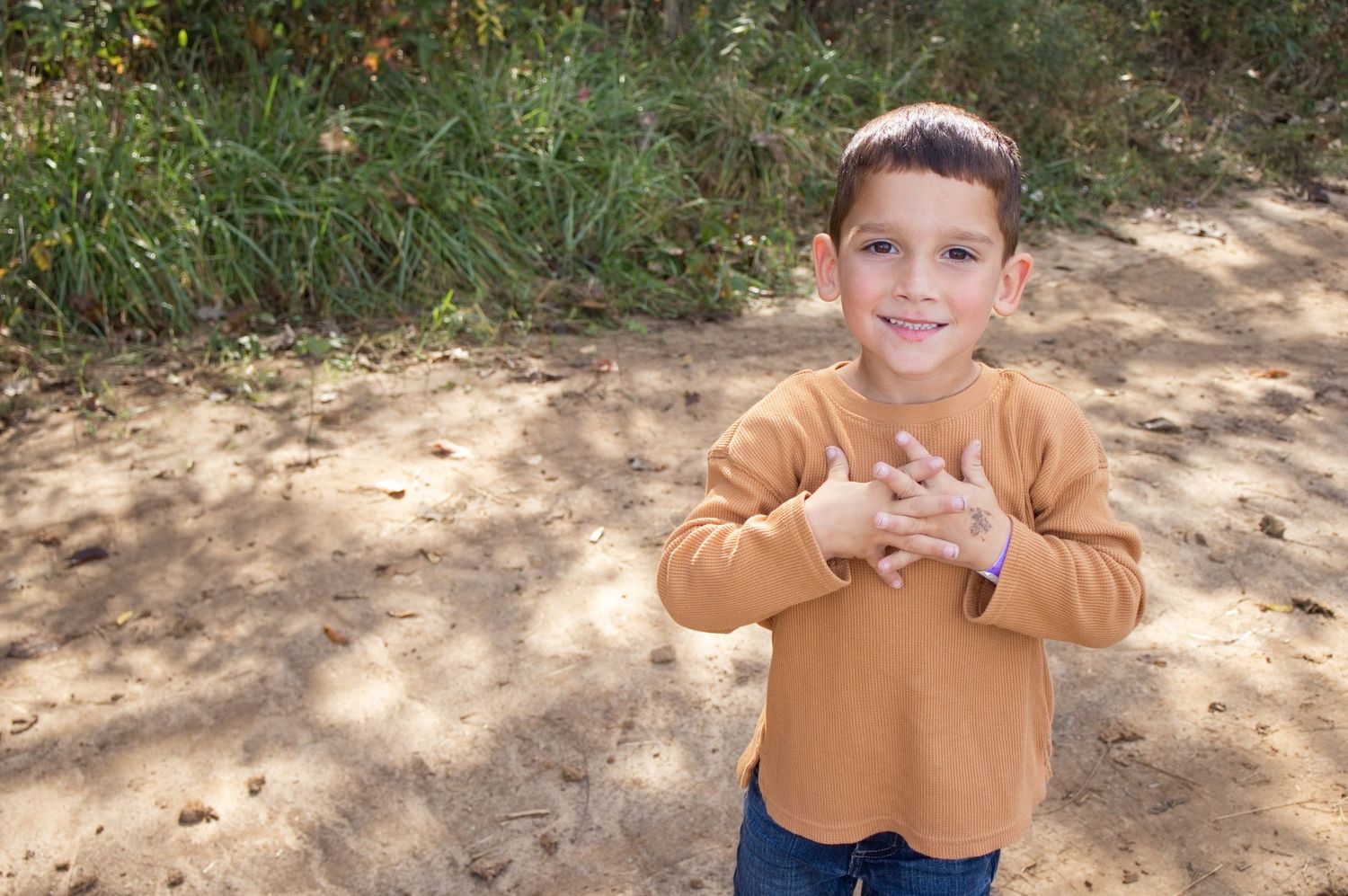 Smiling young boy in an orange sweater and jeans clasps his hands to his chest while standing on a sunlit dirt path near green grass.