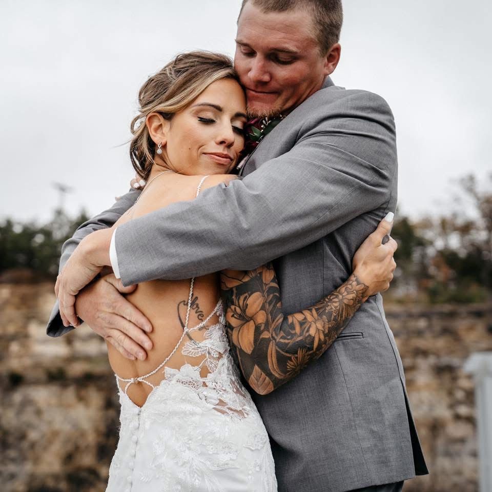 Bride in a lace wedding dress embraces her tattooed groom in a gray suit during an emotional outdoor wedding portrait.