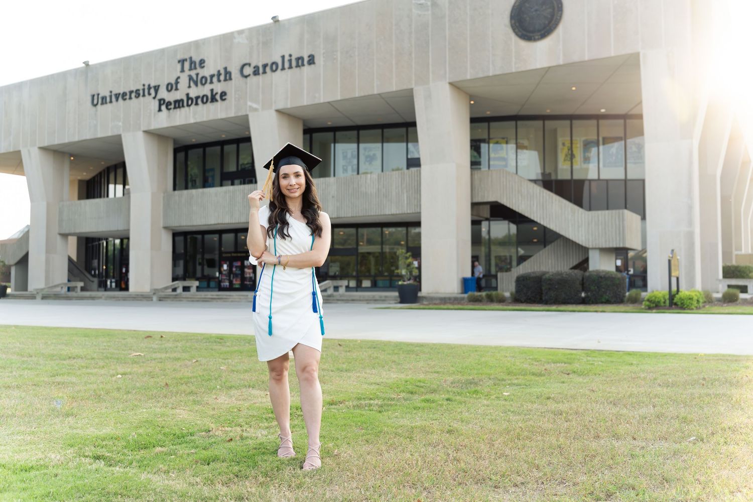 Graduate in cap and gown poses proudly in front of University of North Carolina Pembroke building on sunny day.