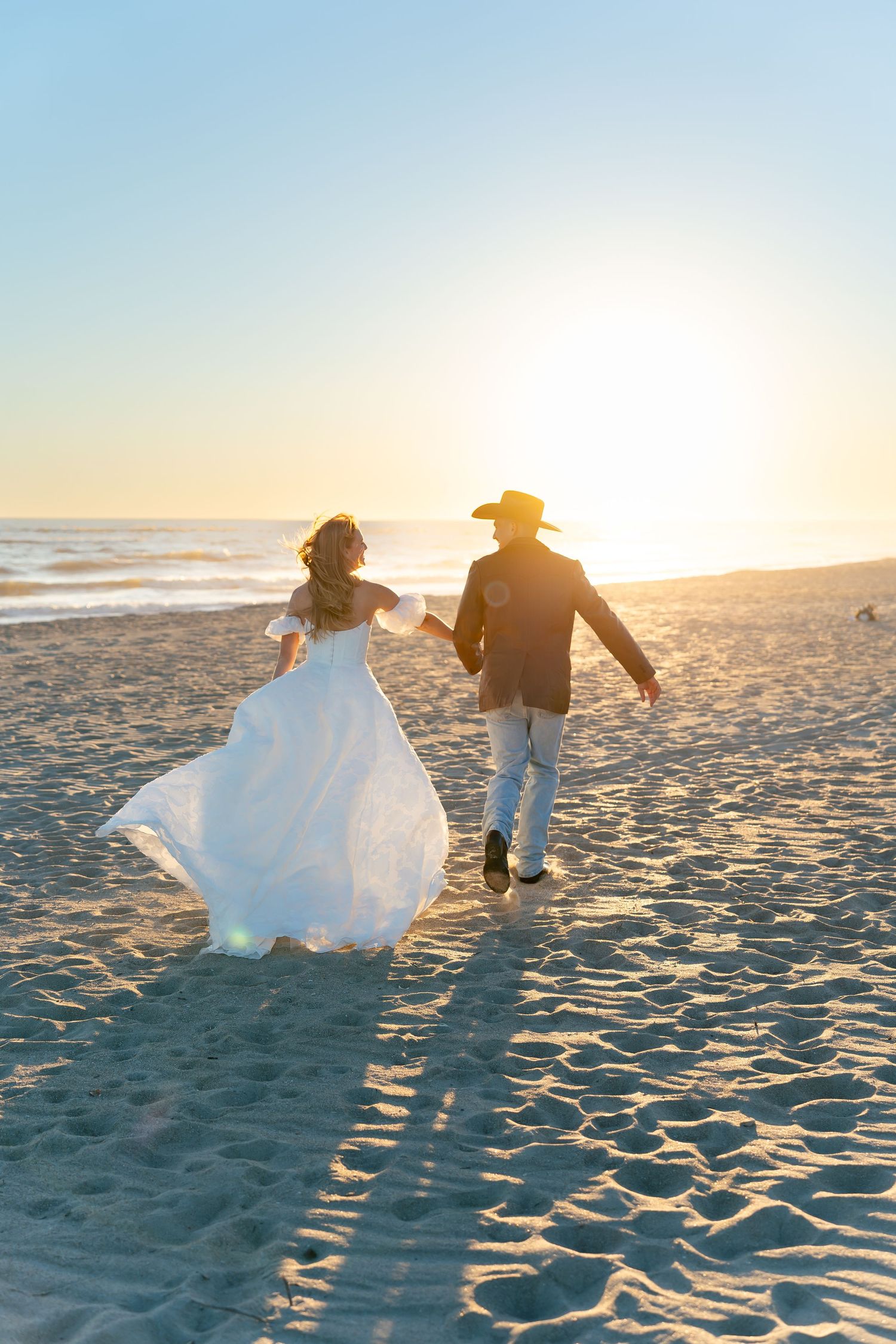 Bride and groom holding hands running on sandy beach at golden sunset, woman in white gown, man in cowboy hat.
