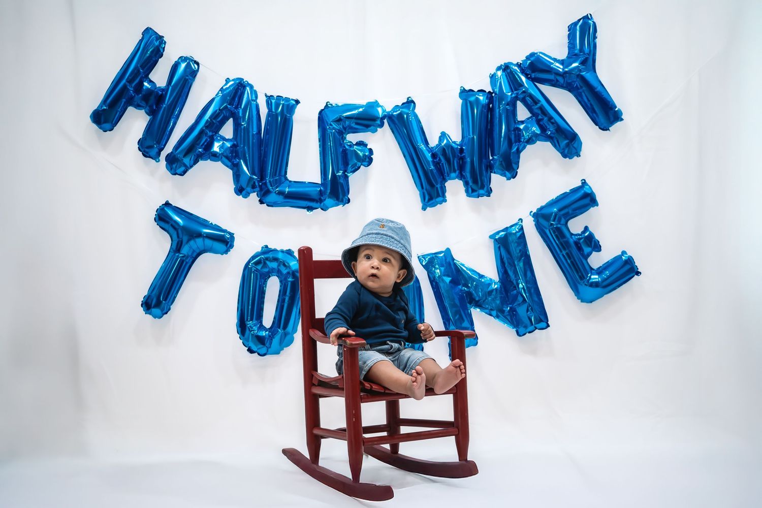 Blue balloon letters spell out 'HALF WAY' above a wooden rocking chair against a white backdrop.