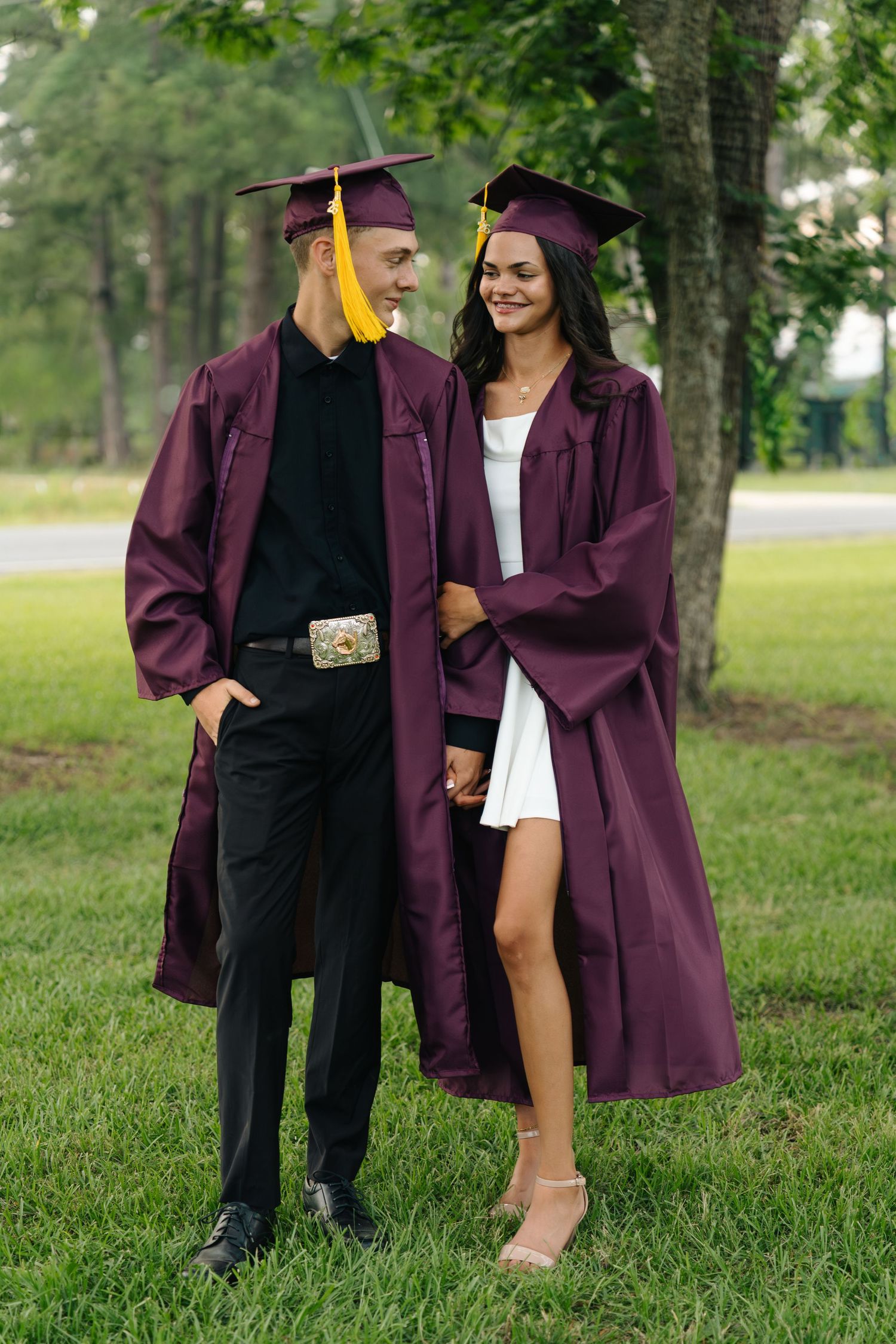 Two graduates in maroon caps and gowns holding hands outdoors on green grass near trees.