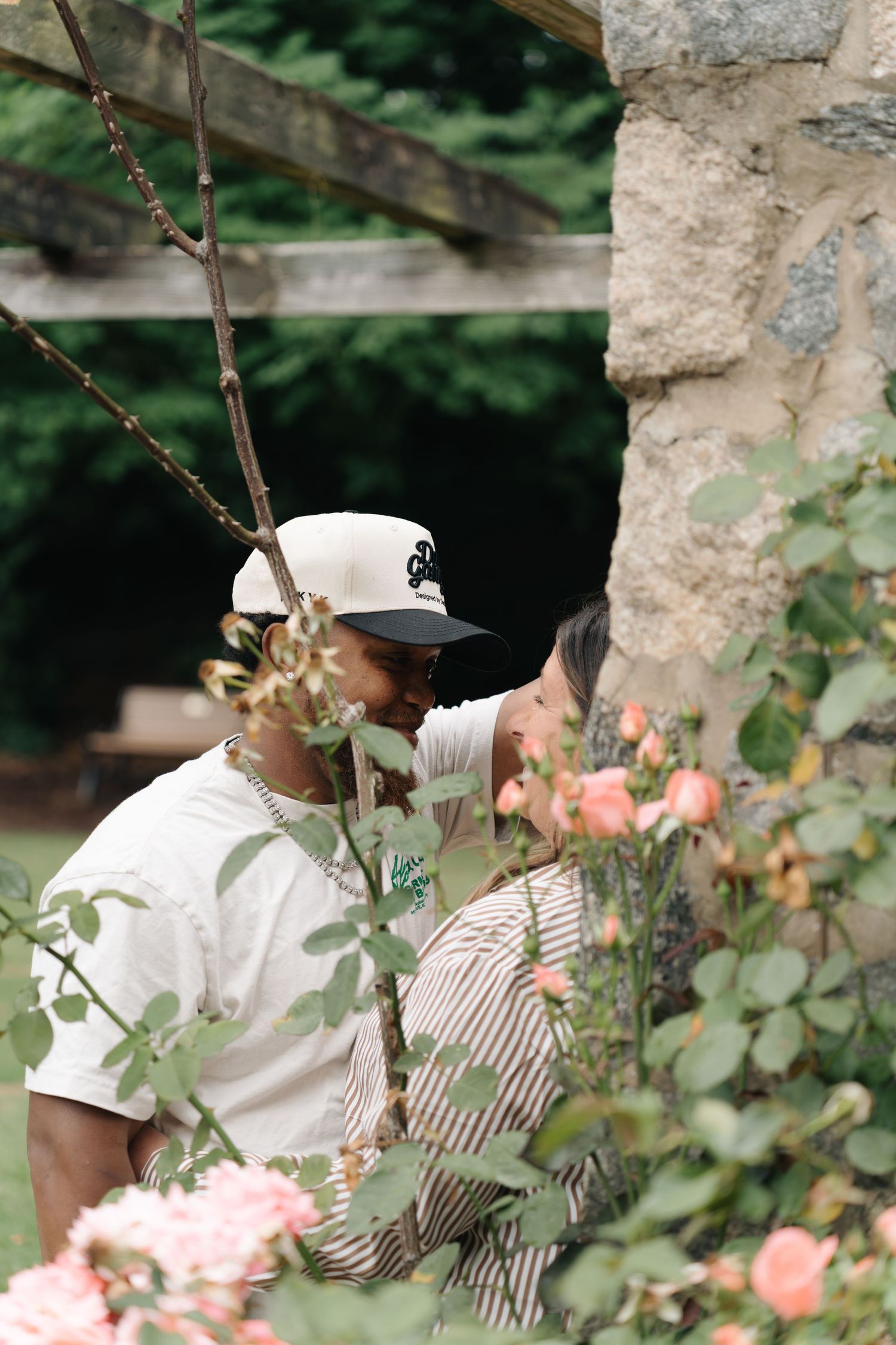 Couple sharing a romantic moment among pink roses and stone wall in a lush garden setting.