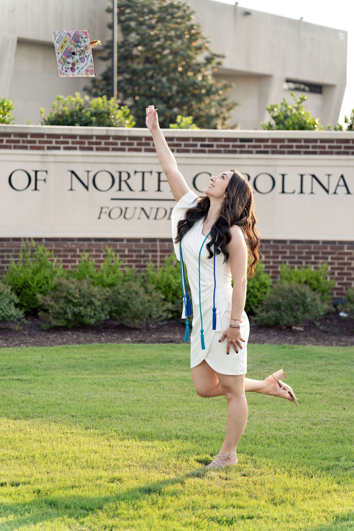 A young woman in a white dress tosses her graduation cap in front of a University of North Carolina sign.