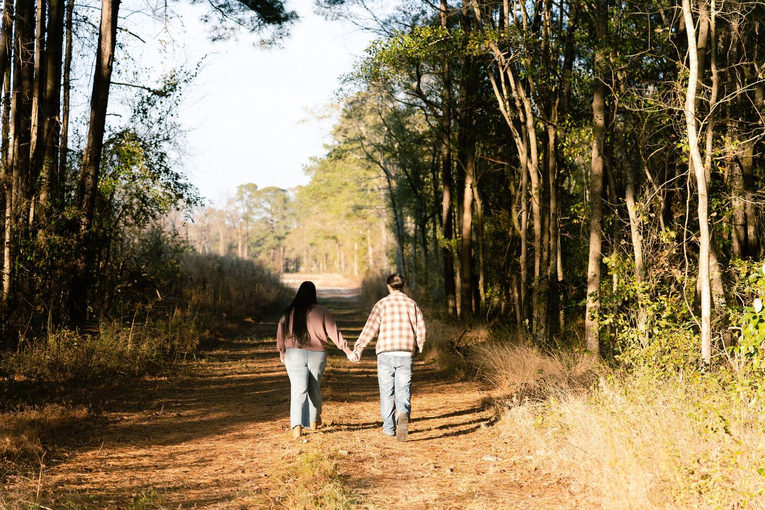 Couple holding hands walking down a sunlit dirt path through a lush green forest on a warm autumn day.