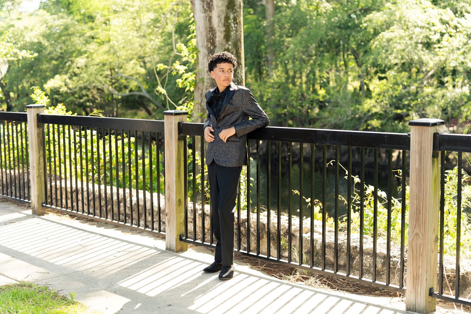 Young man in black suit standing on wooden deck bridge surrounded by lush green trees and water.