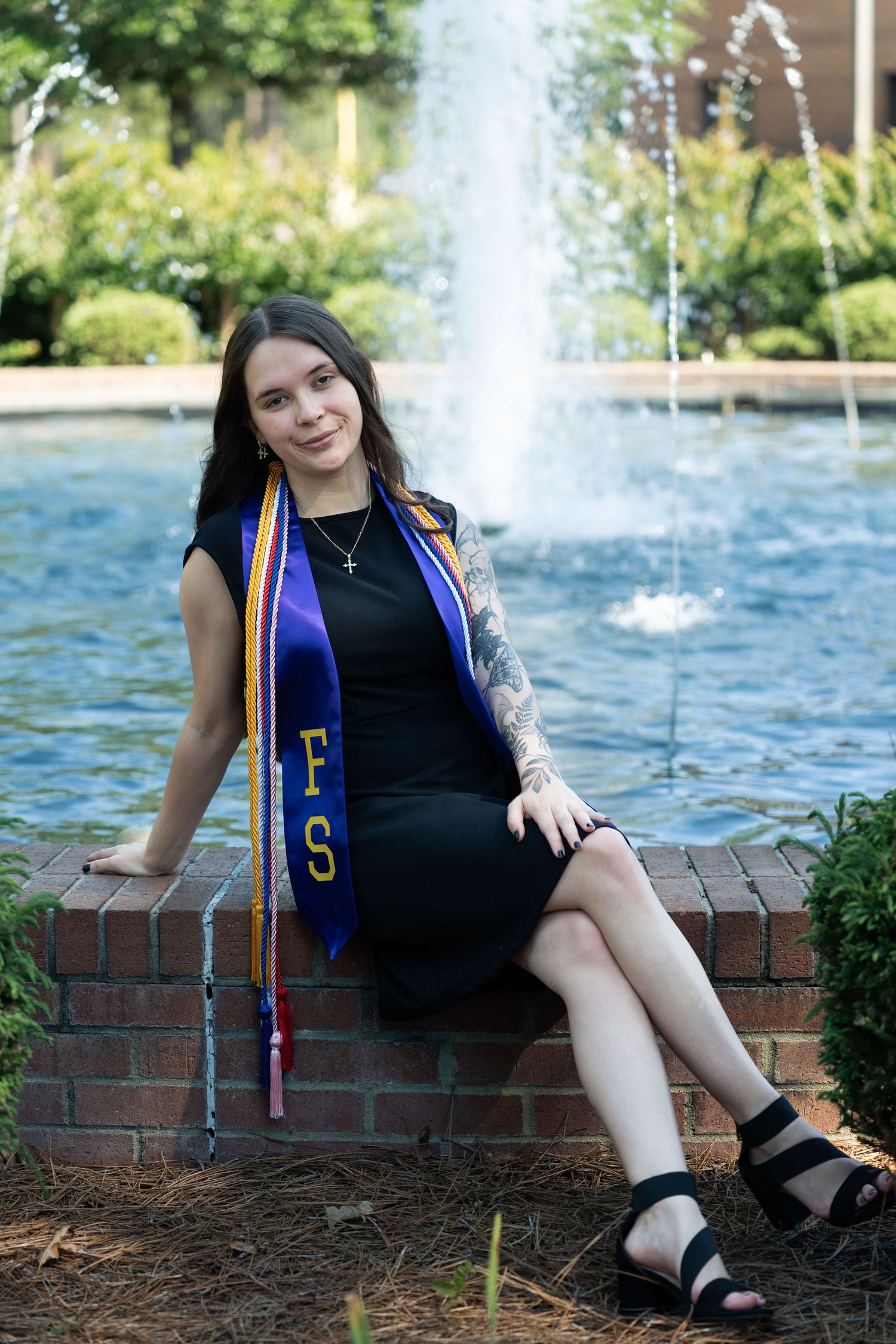 Young woman in black dress with blue graduation stole sits by brick fountain outdoors, celebrating academic achievement.
