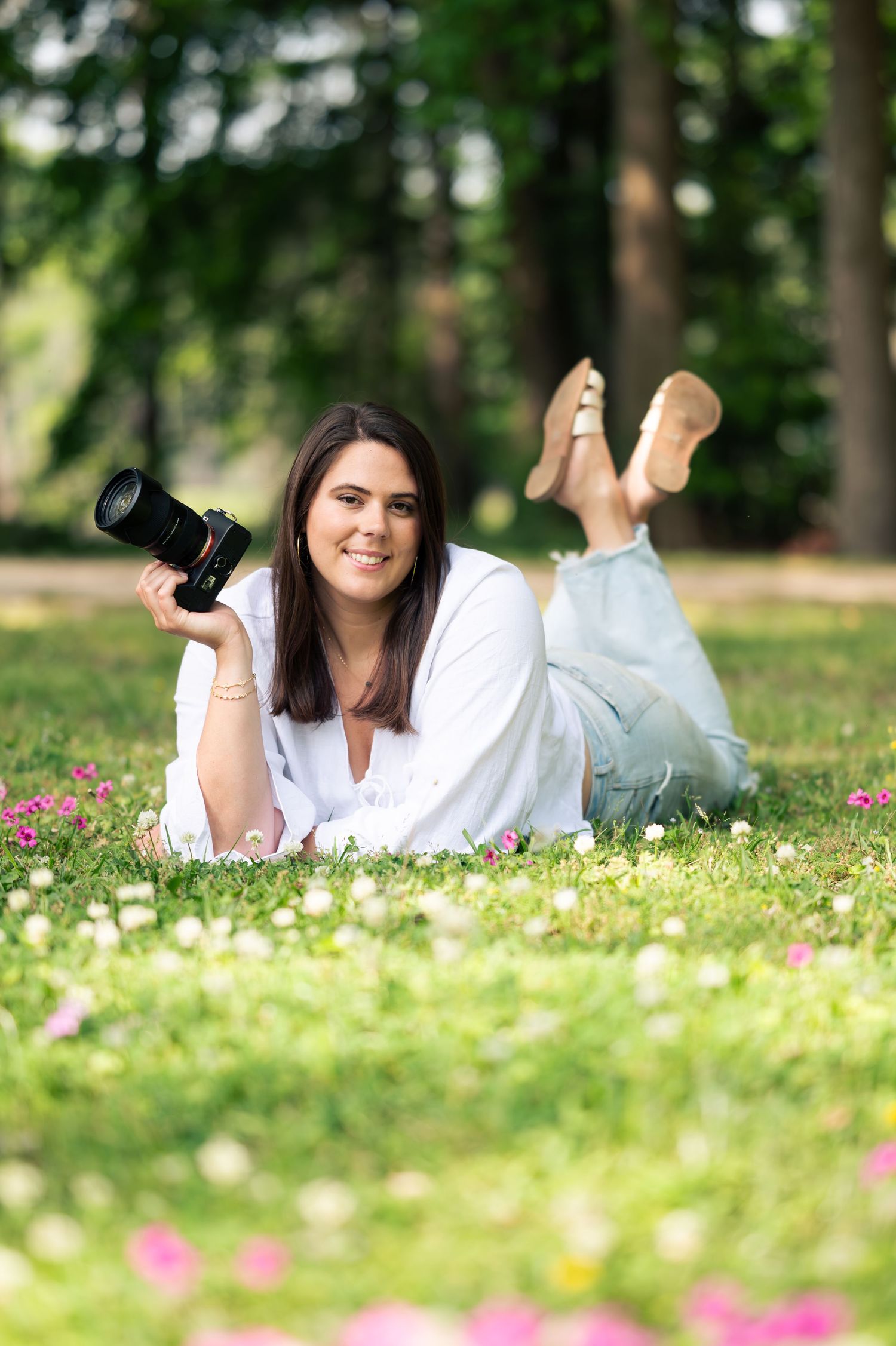 Woman lying on grass in a park, smiling and holding a professional camera, wearing a white blouse and jeans.