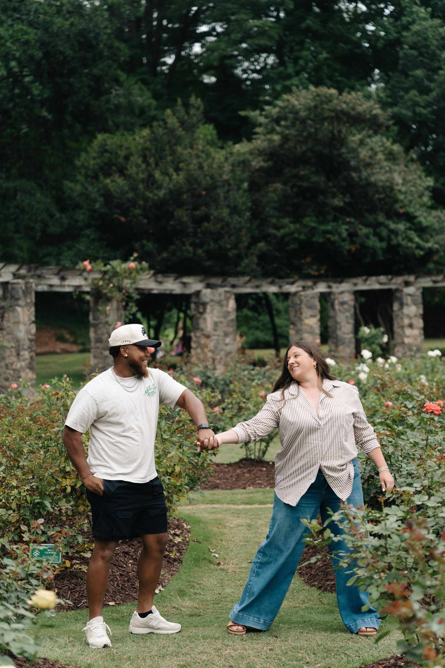 Couple holding hands and smiling in a lush rose garden with stone columns in the background.