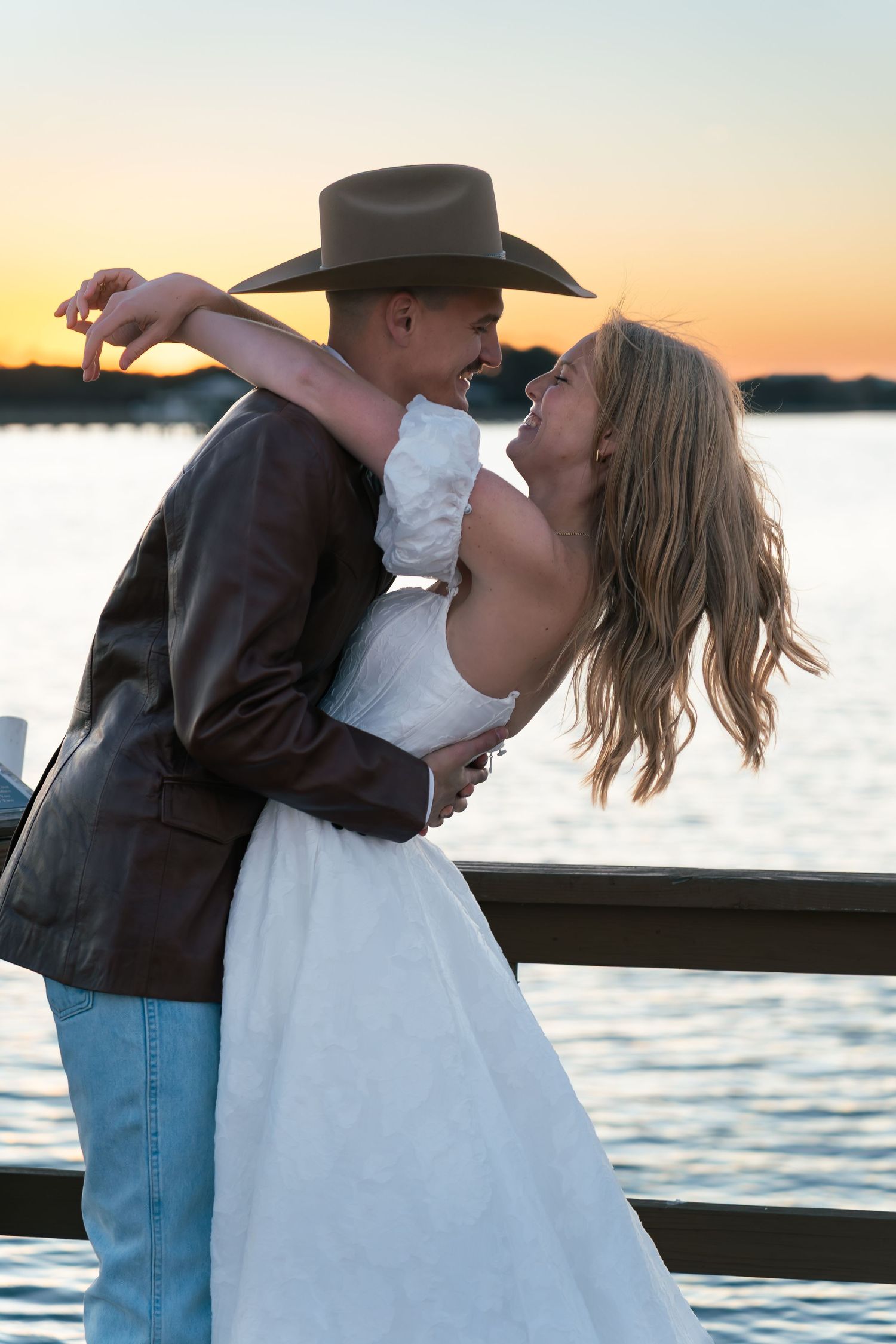 Couple embracing and kissing at sunset by the water, man in cowboy hat and woman in white dress.
