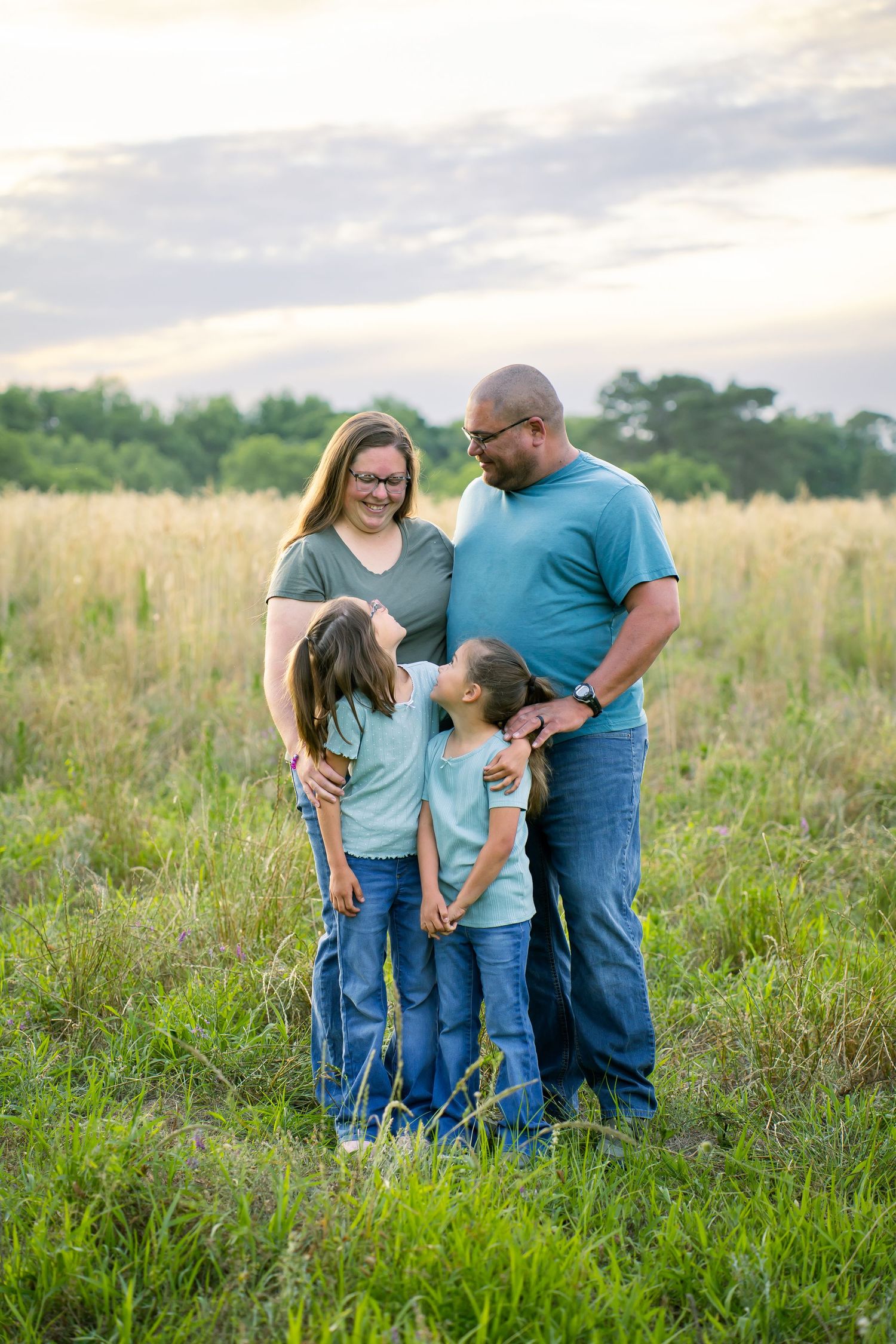 Family wearing coordinating blue outfits stands together in a grassy meadow at sunset.