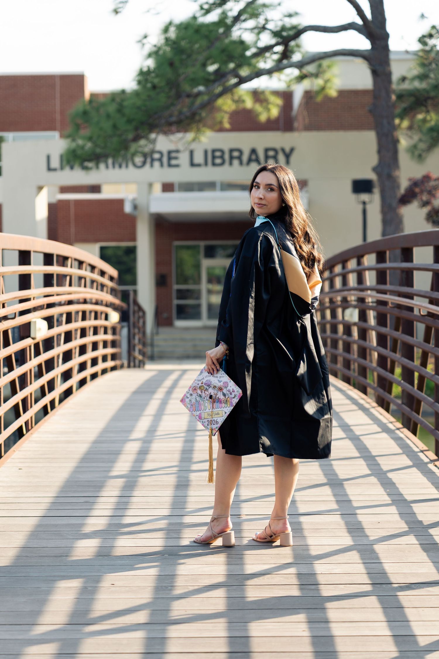 Graduate in black gown holds decorated cap on wooden bridge outside Livermore Library.