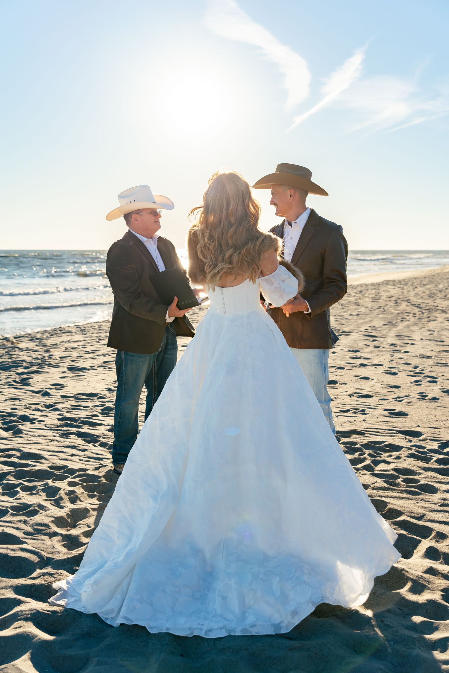 Bride in white gown with two men in cowboy hats exchanging vows on a sunny beach at sunset.