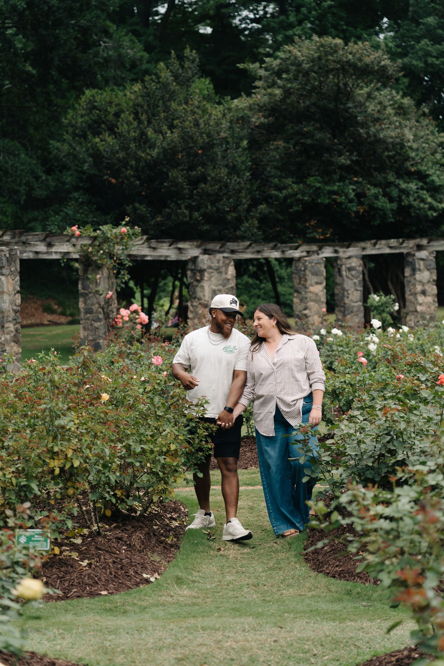 Couple walking hand in hand through a lush rose garden with stone columns in the background.
