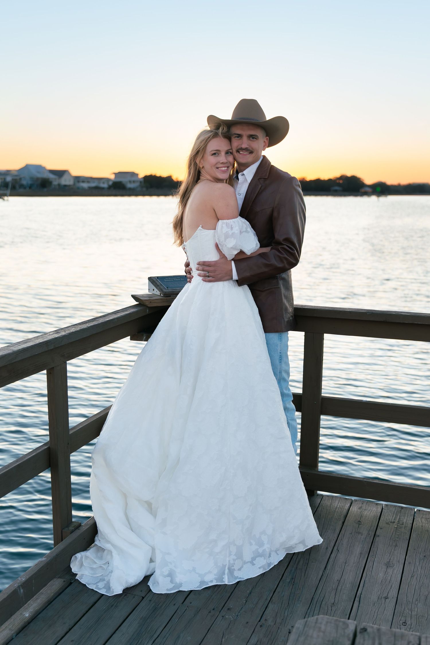 Bride and groom embrace on a wooden dock at sunset, she in a white gown, he in a brown suit and cowboy hat.