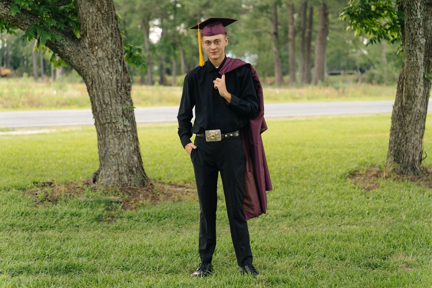 Young man in maroon graduation cap and gown posing outdoors on green grass near trees for graduation photos.