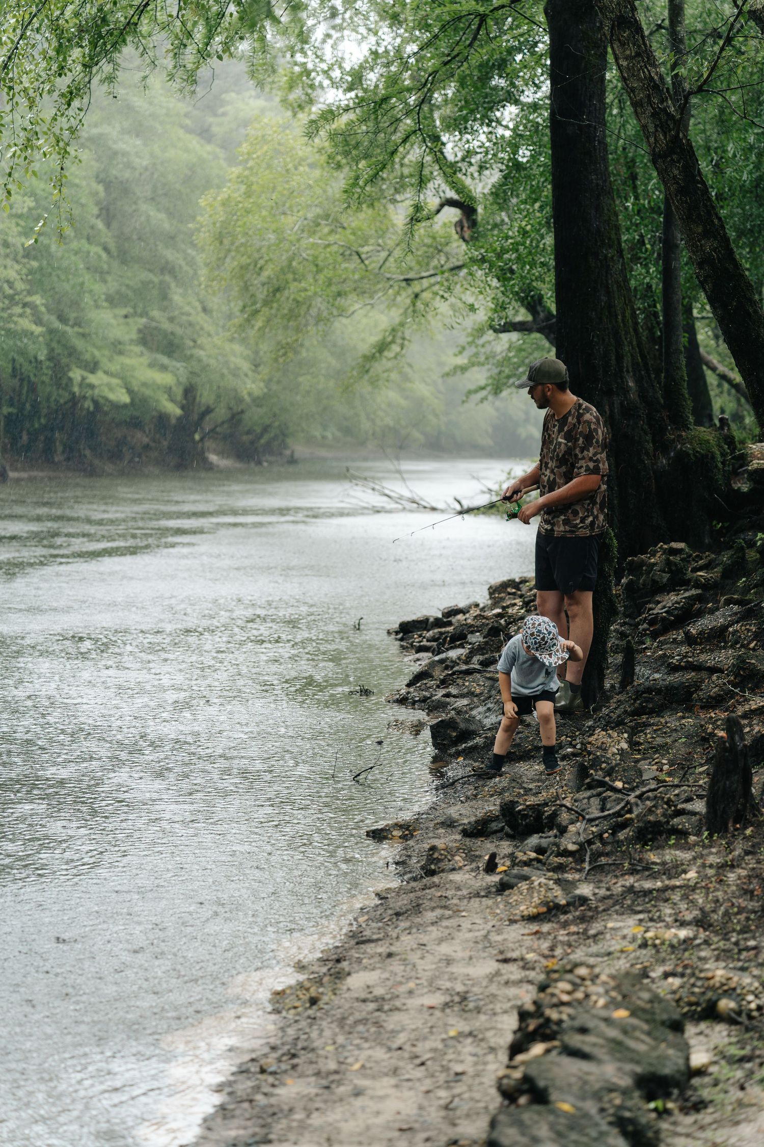 Adult and toddler fishing together on a misty river bank surrounded by lush green trees.
