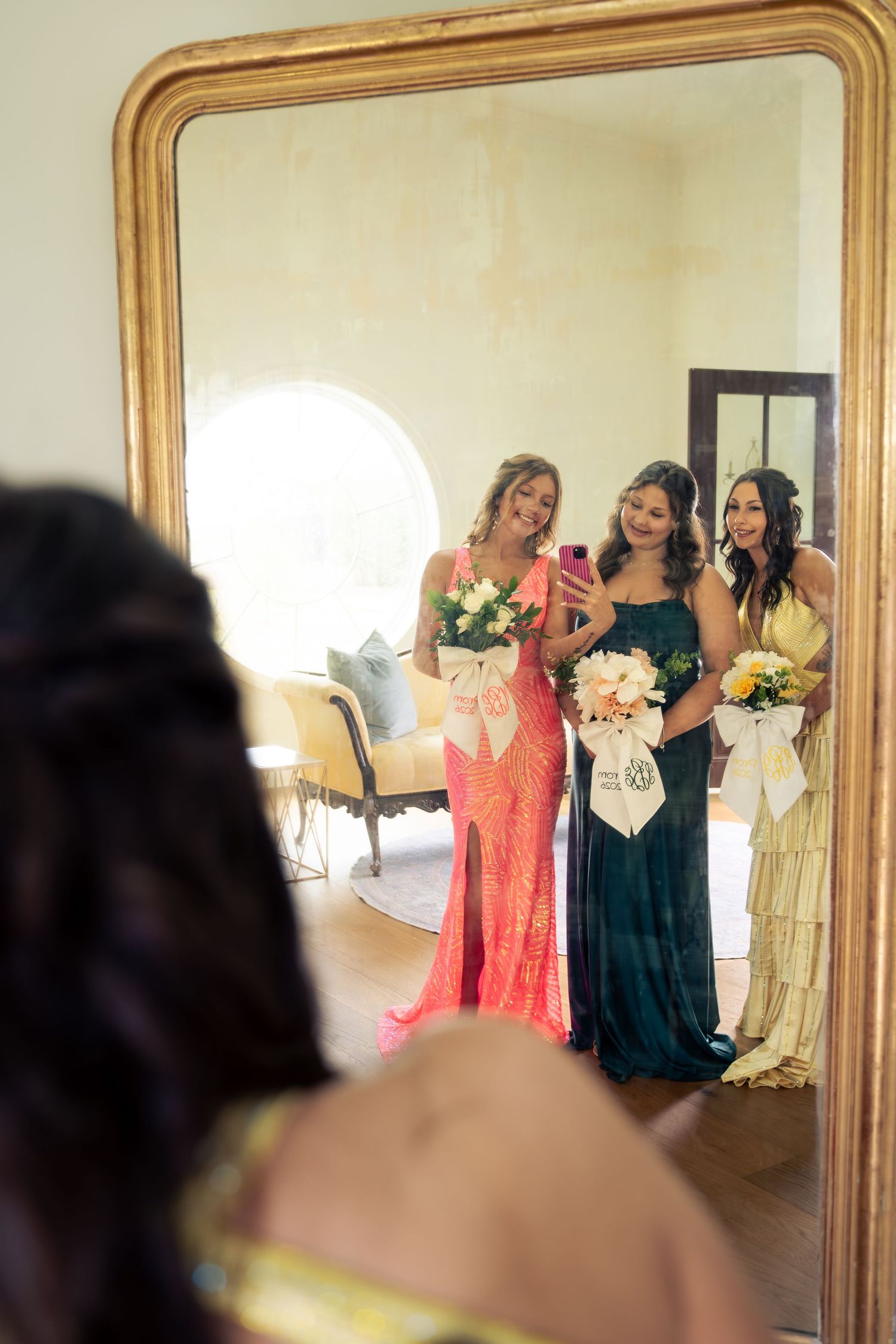 Three women in formal gowns take a mirror selfie, holding bouquets, with one wearing a bridal sash.