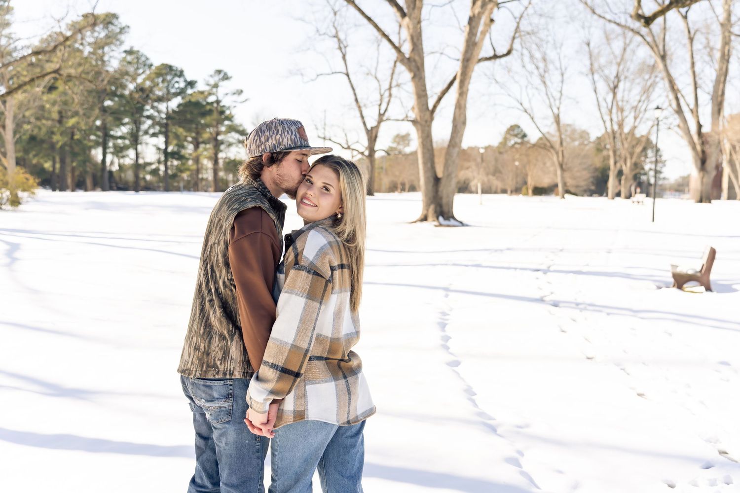 Couple embracing in snowy park, wearing flannel and denim, surrounded by snow-covered ground and bare trees.