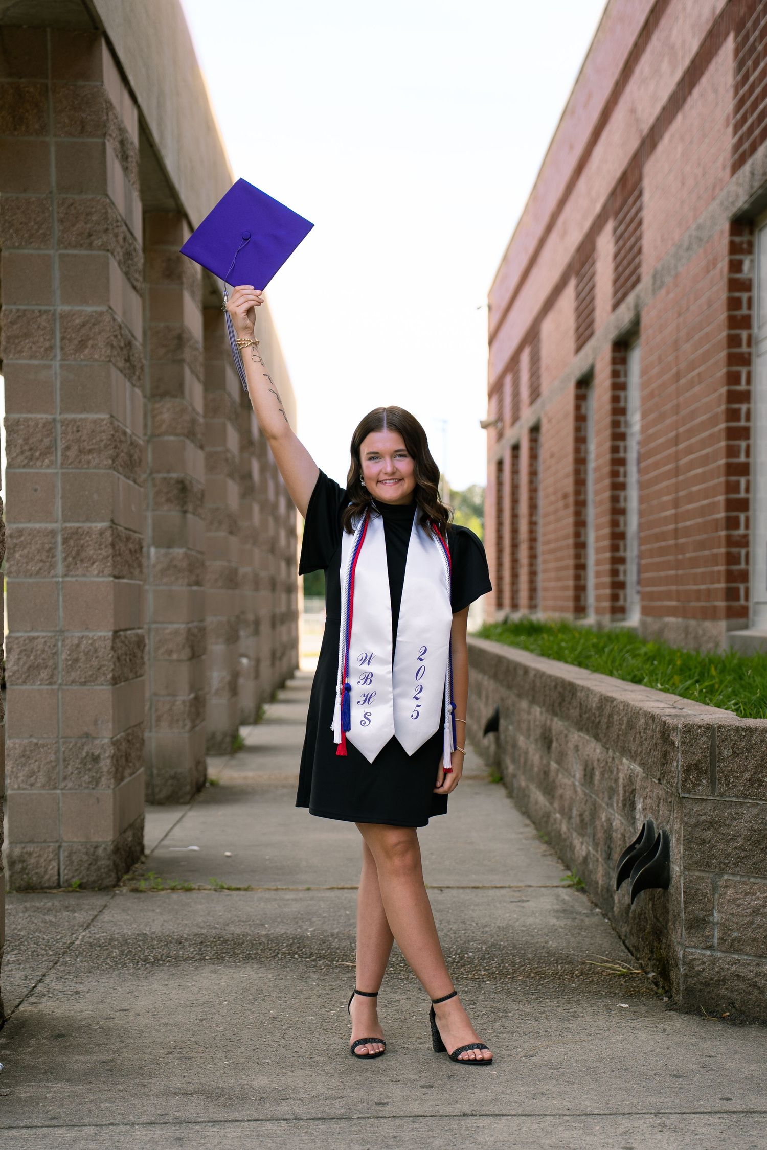 Graduate in cap and gown poses between stone columns on campus walkway.