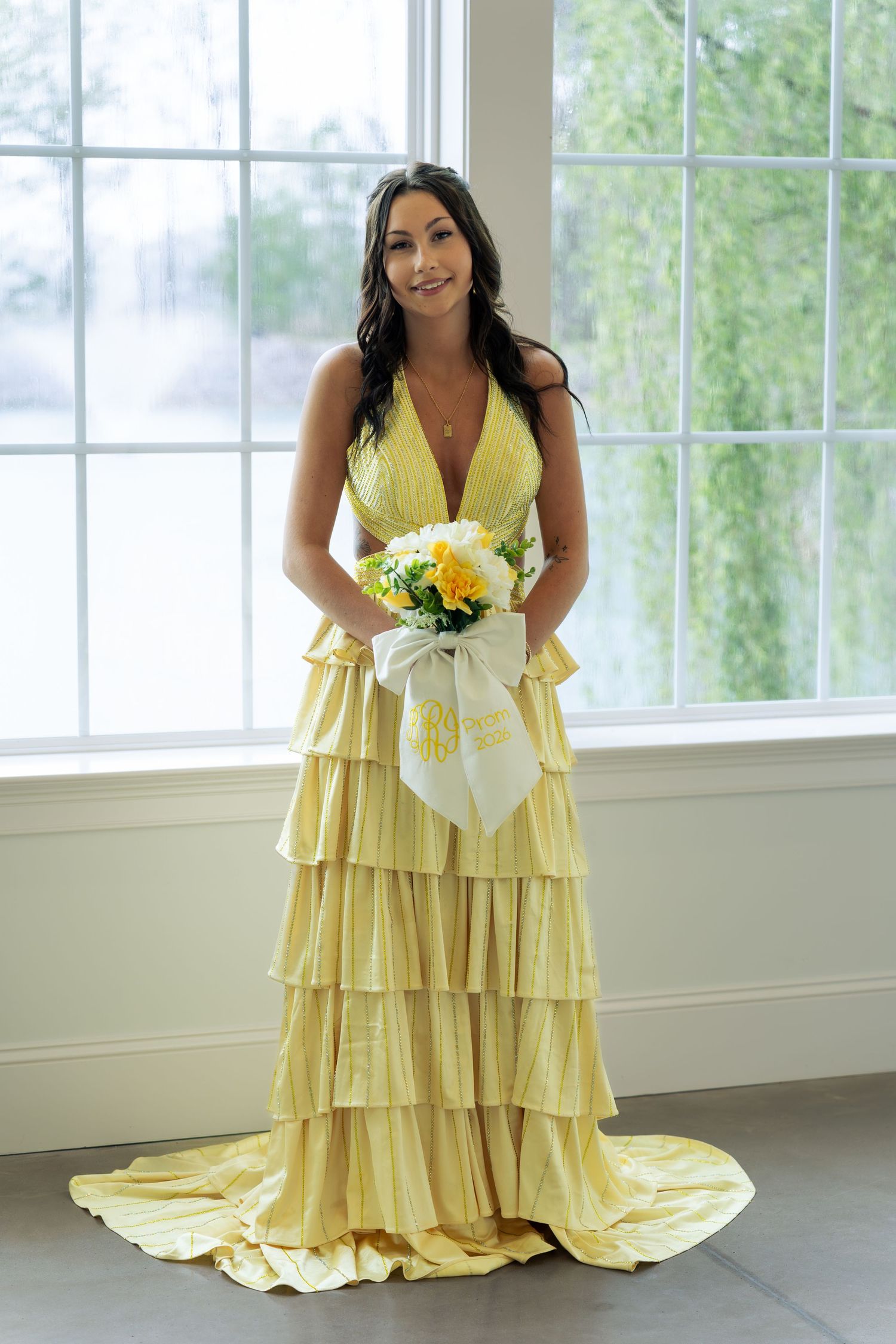 Woman in yellow tiered halter gown holding yellow and white bouquet standing by large window.