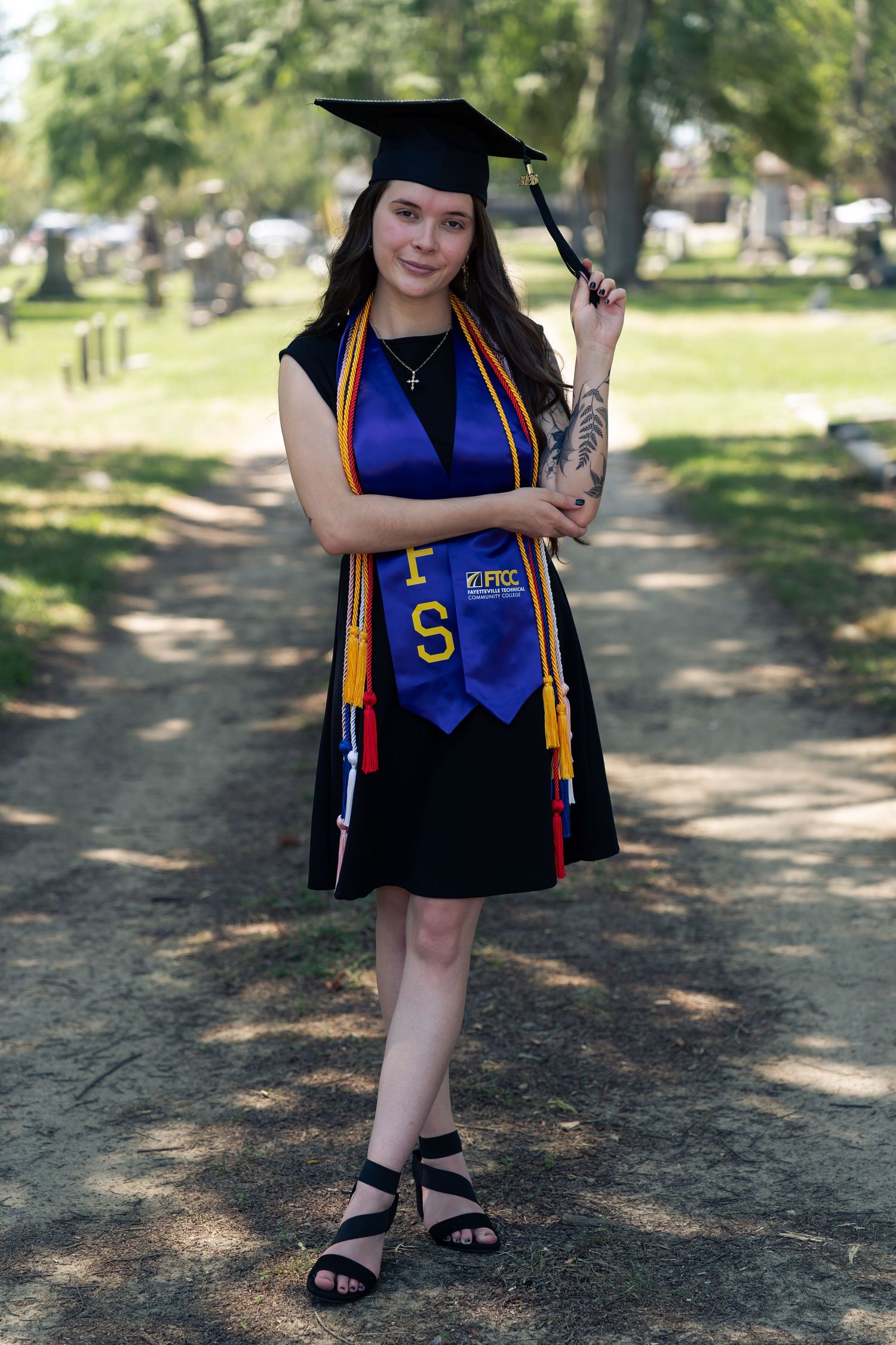 Young woman in black graduation gown, cap, and blue FRC stole poses outdoors holding tassel, celebrating academic achievement.