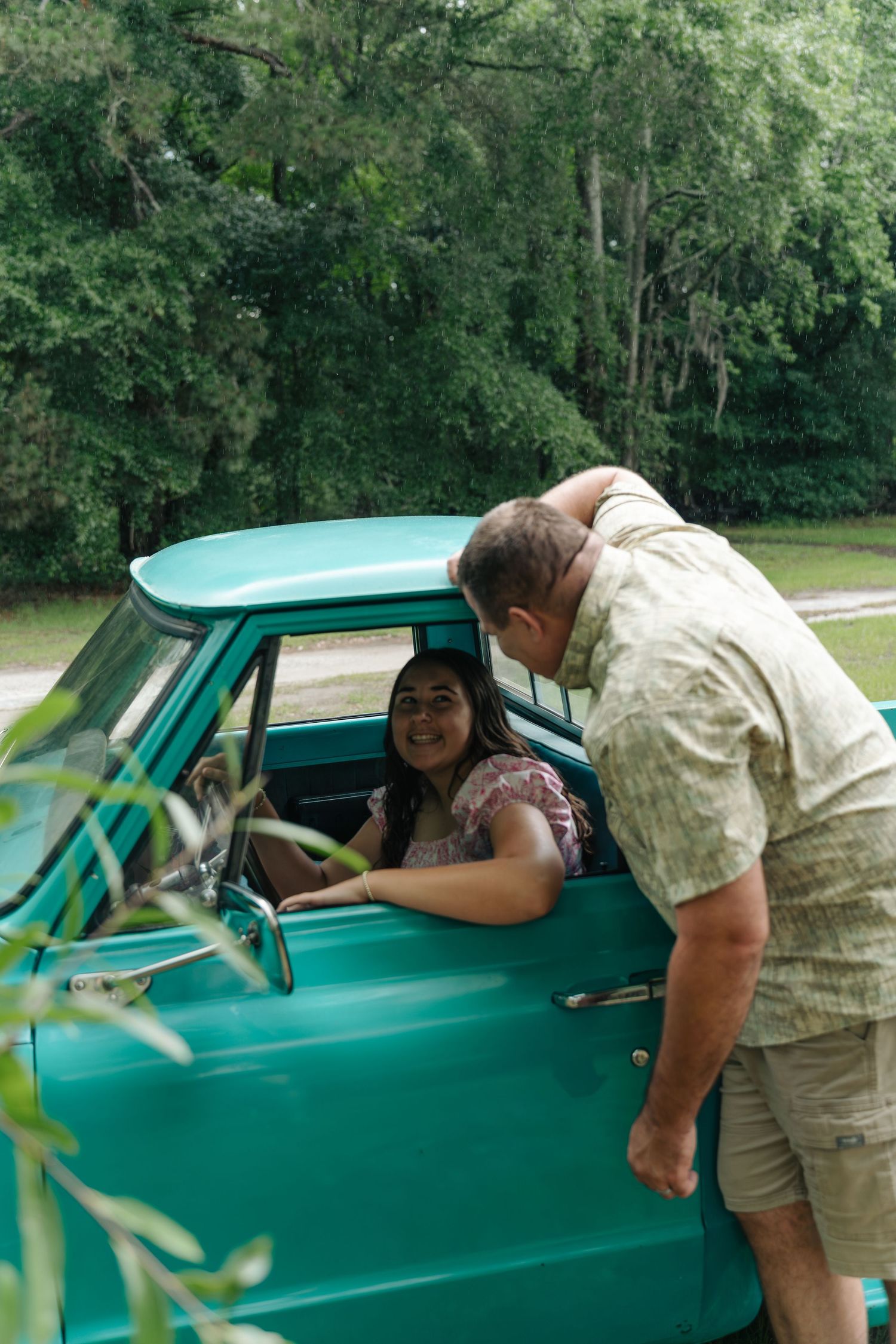 A woman sits in a vintage teal pickup truck smiling at a man leaning on the door outdoors.