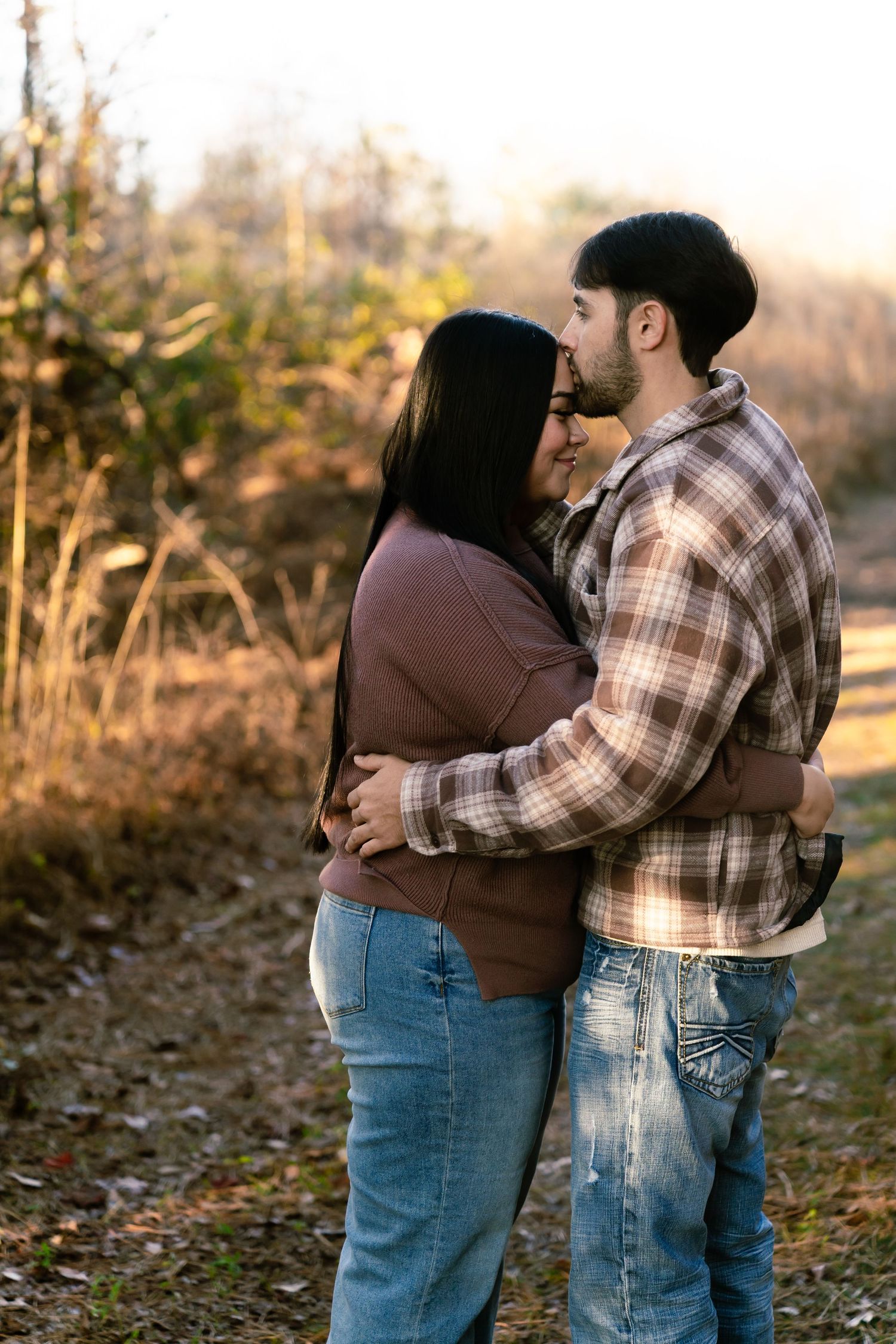 Couple embracing outdoors in autumn, man kissing woman's cheek, both wearing jeans and casual tops, golden fall foliage background.