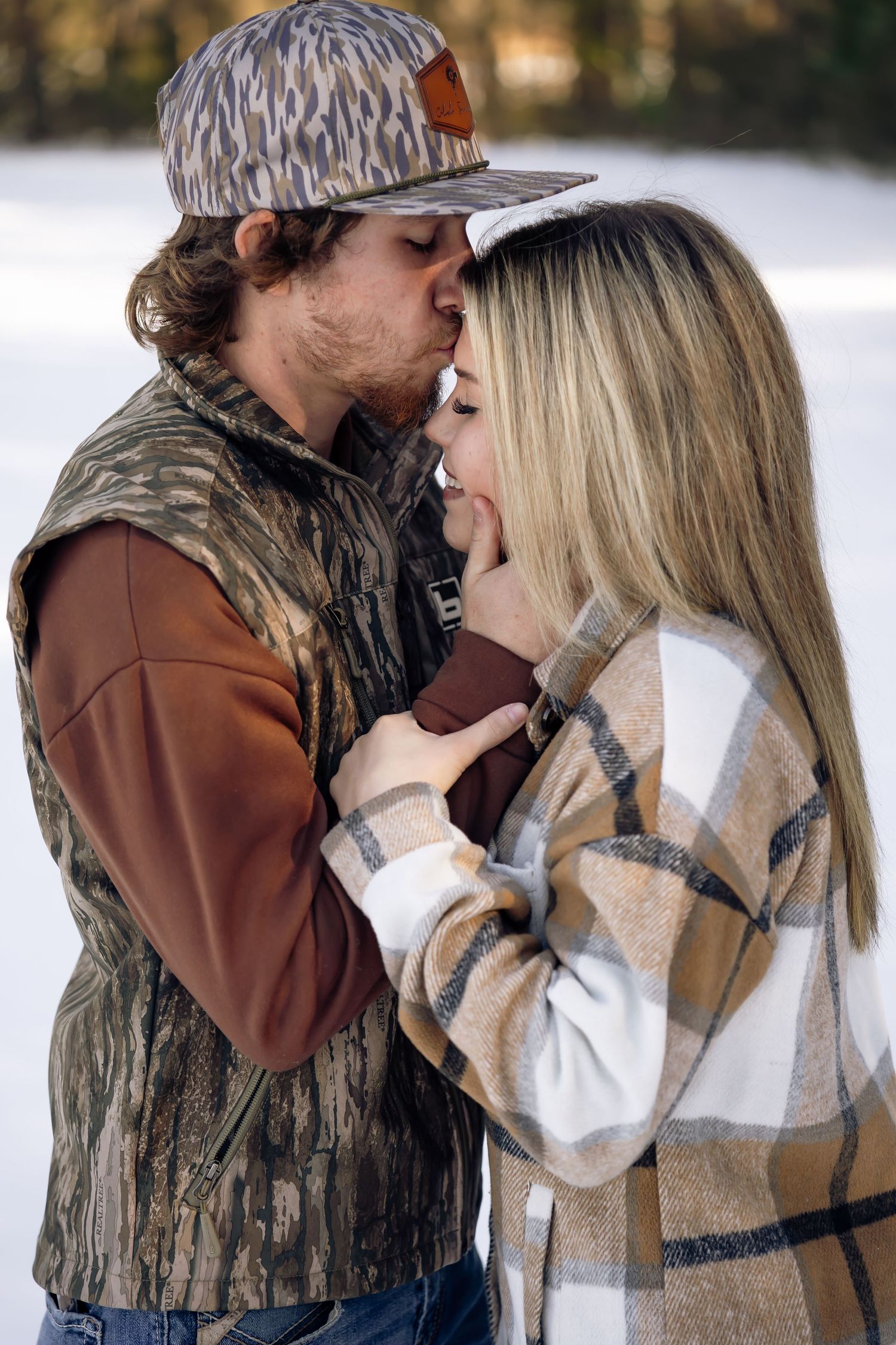 Couple sharing a romantic kiss outdoors in a snowy winter setting, wearing casual country-style outfits.