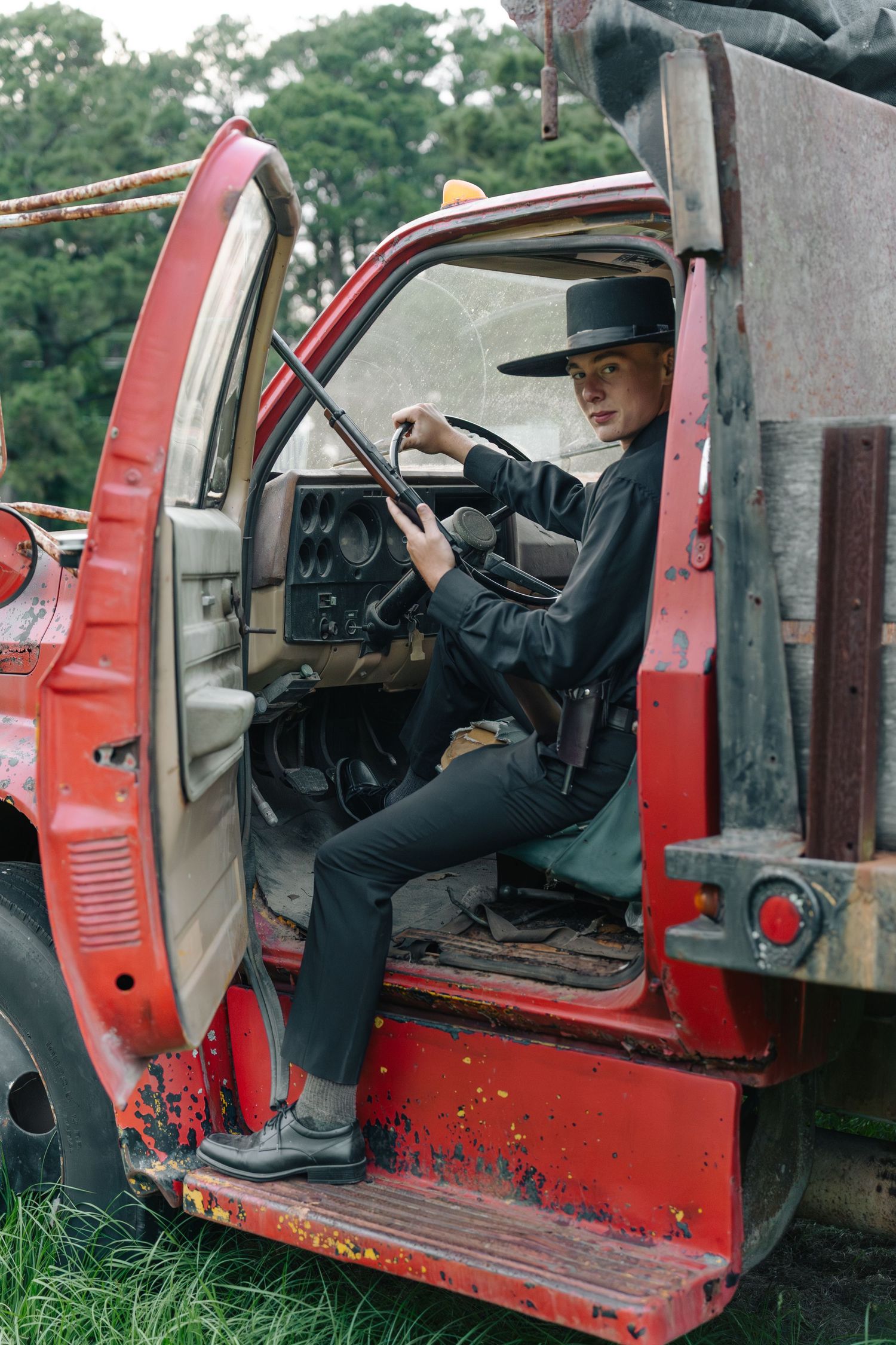 Young person in black outfit and hat sitting in driver seat of vintage red truck outdoors.