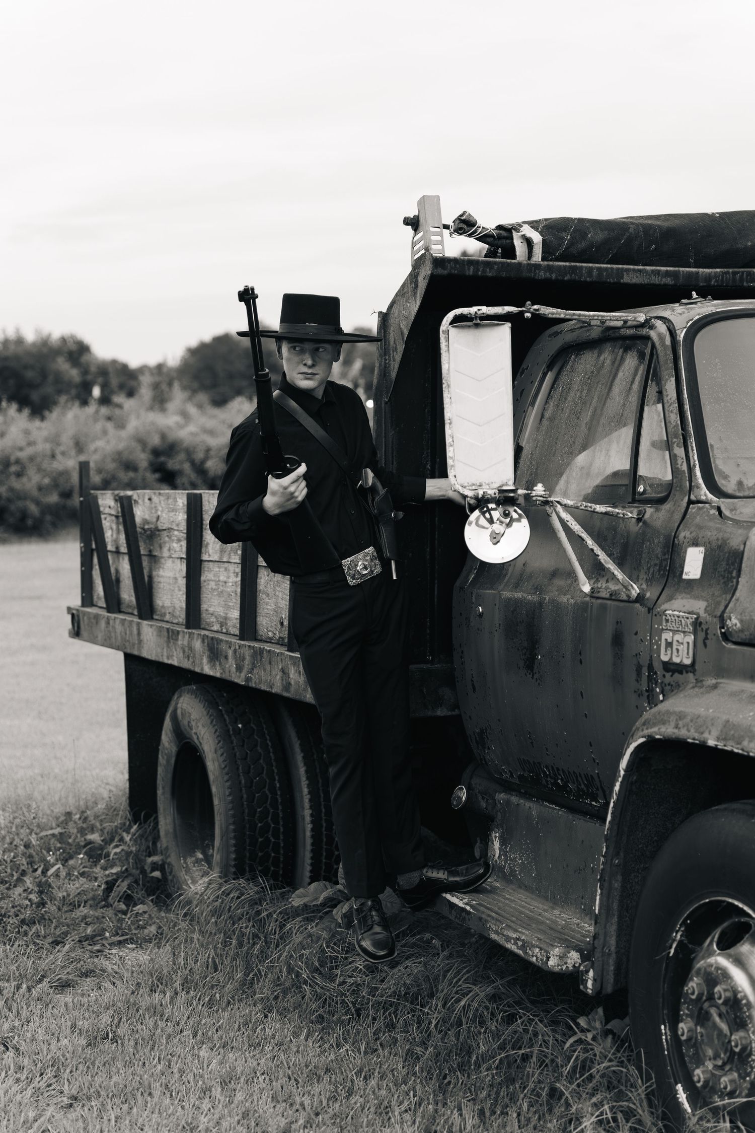 Man in black outfit and cowboy hat holding rifle beside vintage truck in black and white photo.