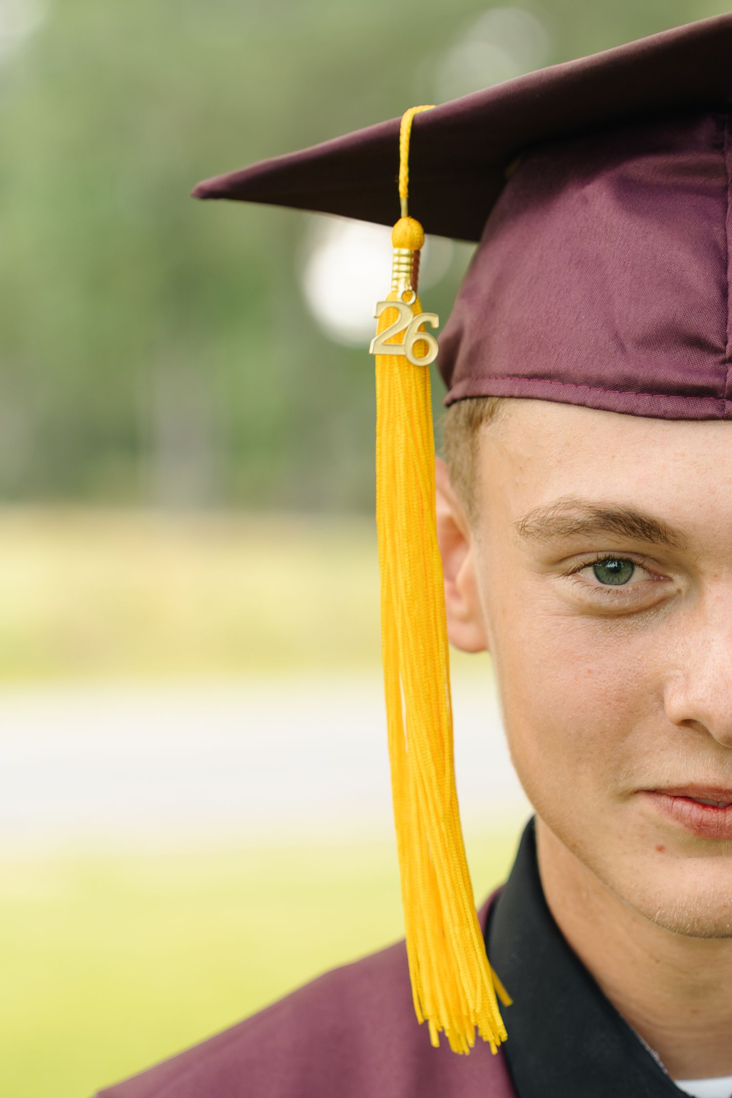 Young male graduate in maroon cap and gown with gold tassel and 26 charm poses outdoors for graduation portrait.