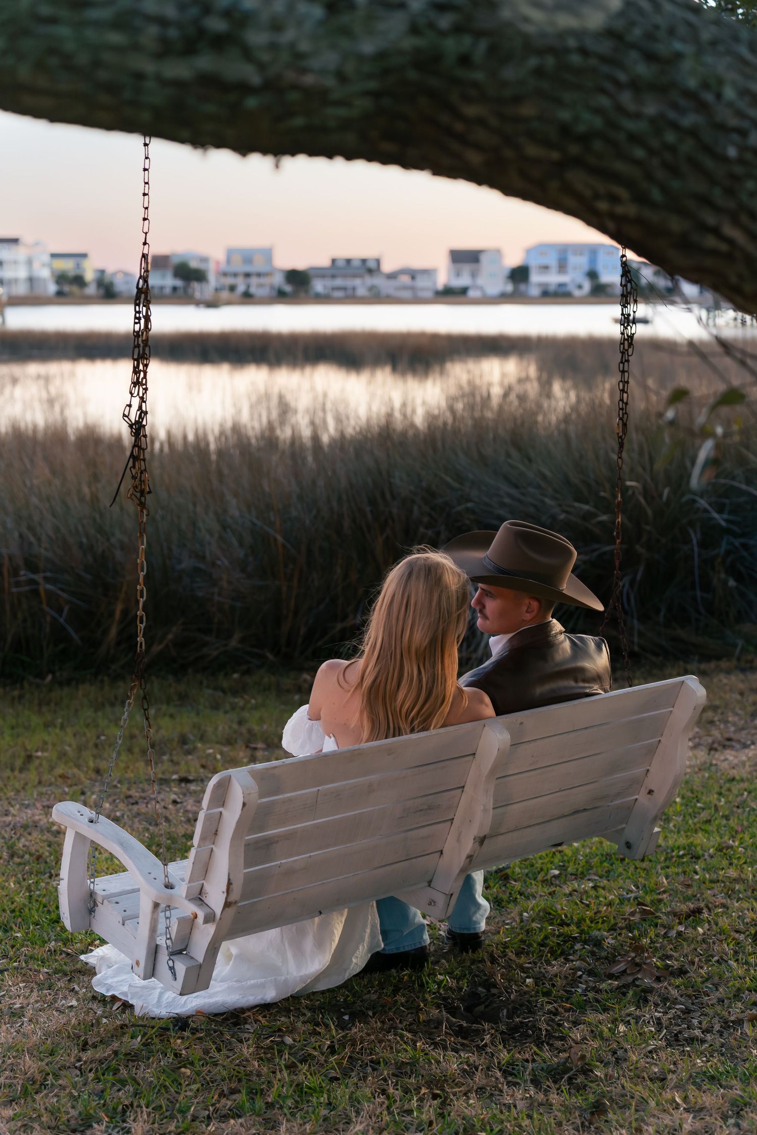 Couple in wedding attire sitting on a white swing bench by a marsh at sunset, groom wearing a cowboy hat.