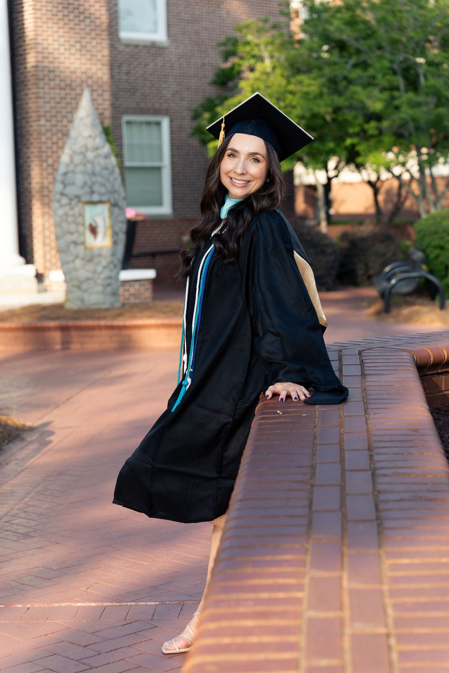A smiling woman in black graduation cap and gown poses outdoors on a brick pathway at her college campus.