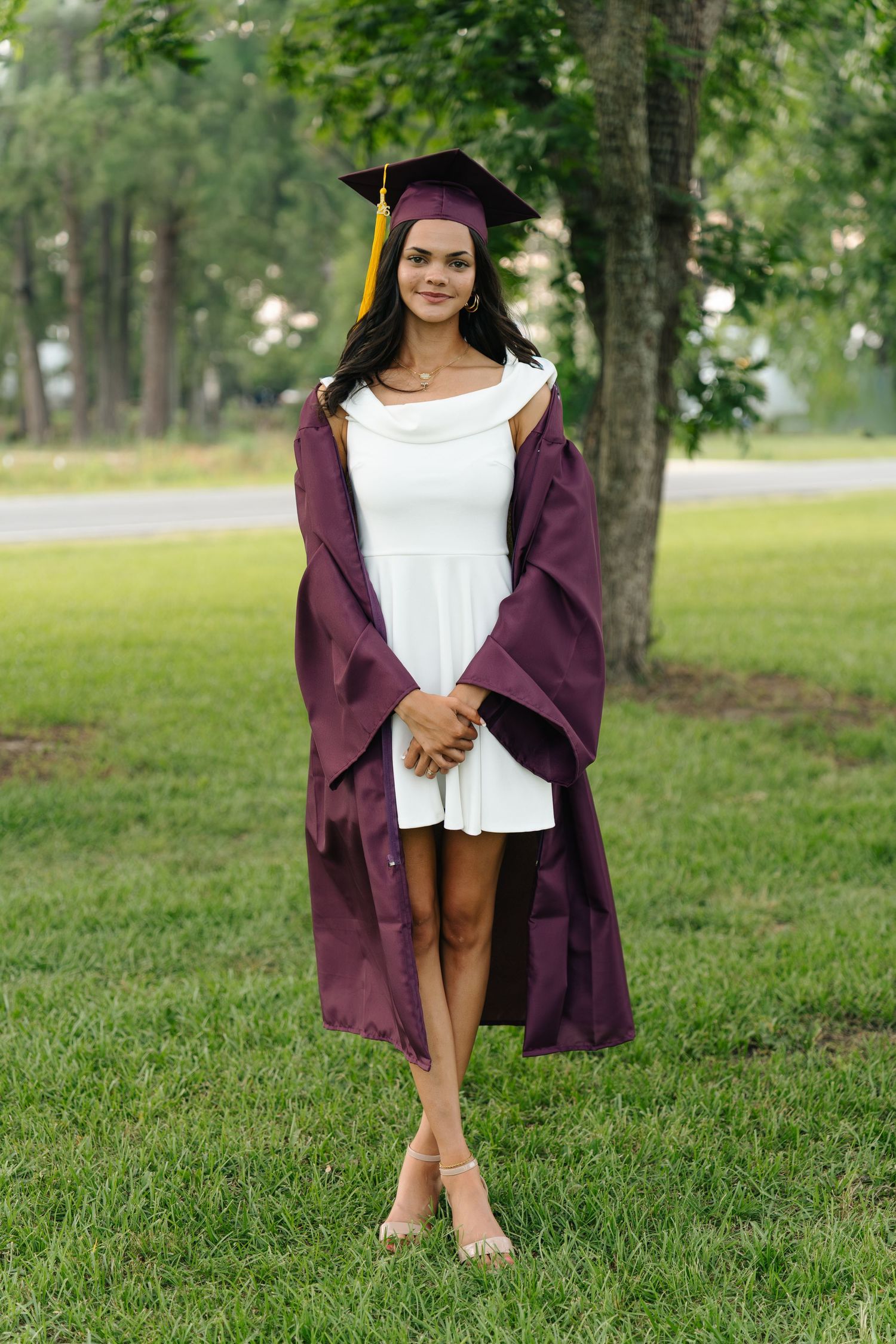 Young woman in maroon graduation cap and gown over white dress, posing outdoors on green grass near trees.