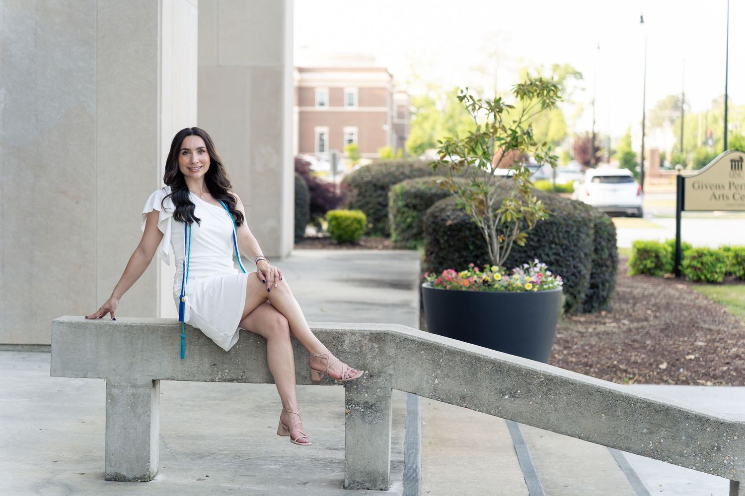 Young woman in white dress sitting on concrete bench outdoors near modern building with greenery.
