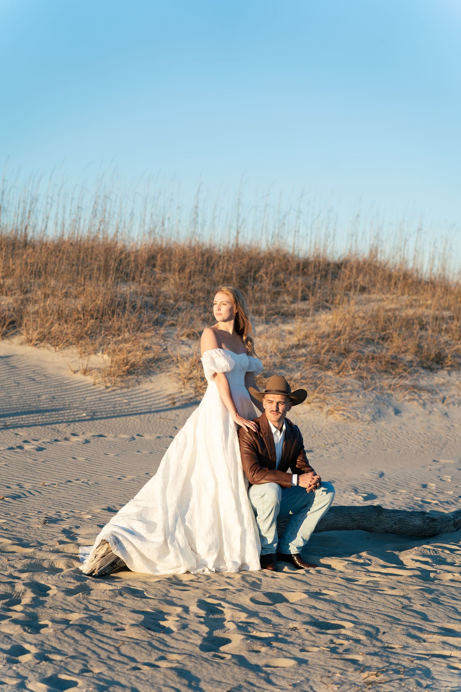 Bride in white off-shoulder gown stands behind groom in cowboy hat on sandy beach dunes at golden hour.