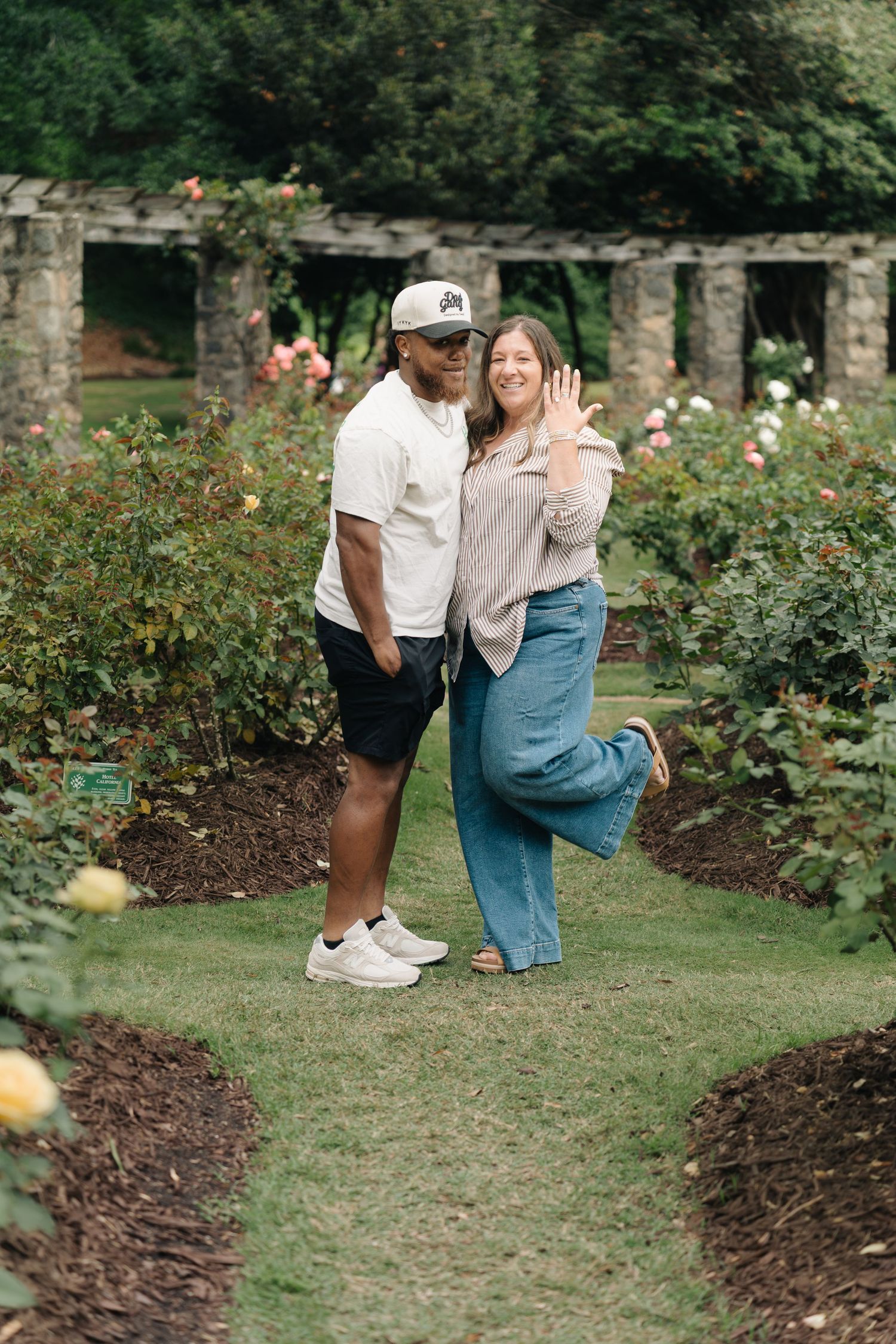Happy couple posing in a rose garden, woman showing engagement ring, surrounded by lush greenery and stone pergola.