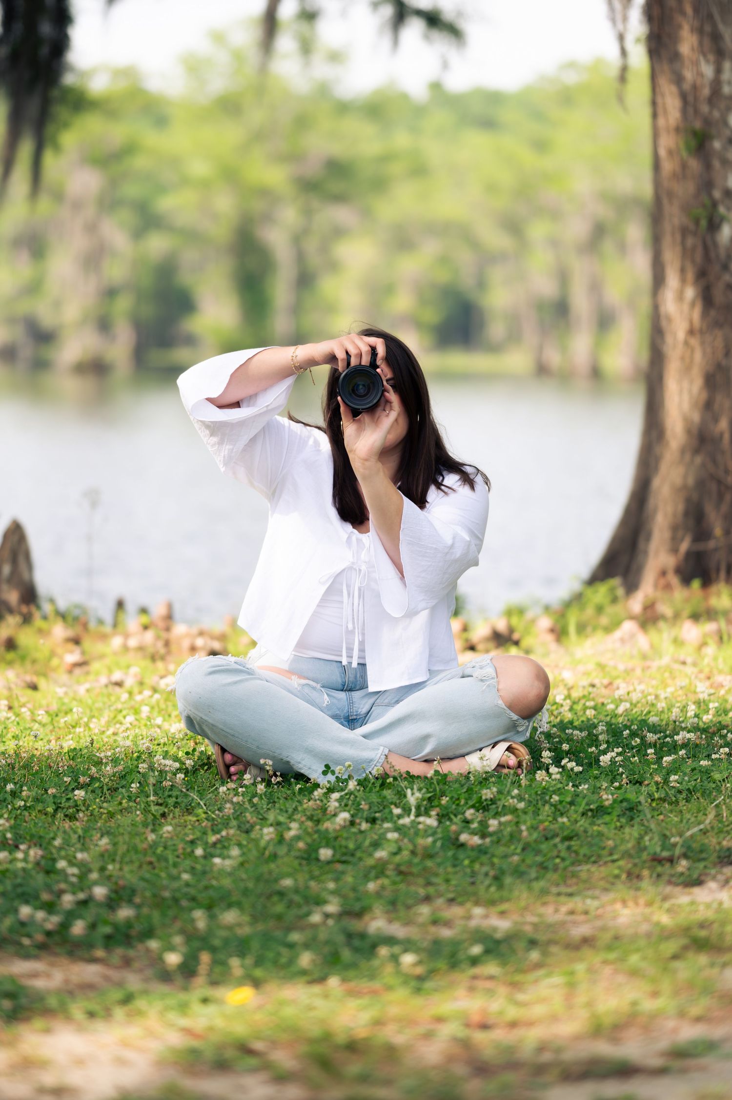 Woman in white shirt sitting cross-legged on grass, taking photo with DSLR camera in a serene park setting.