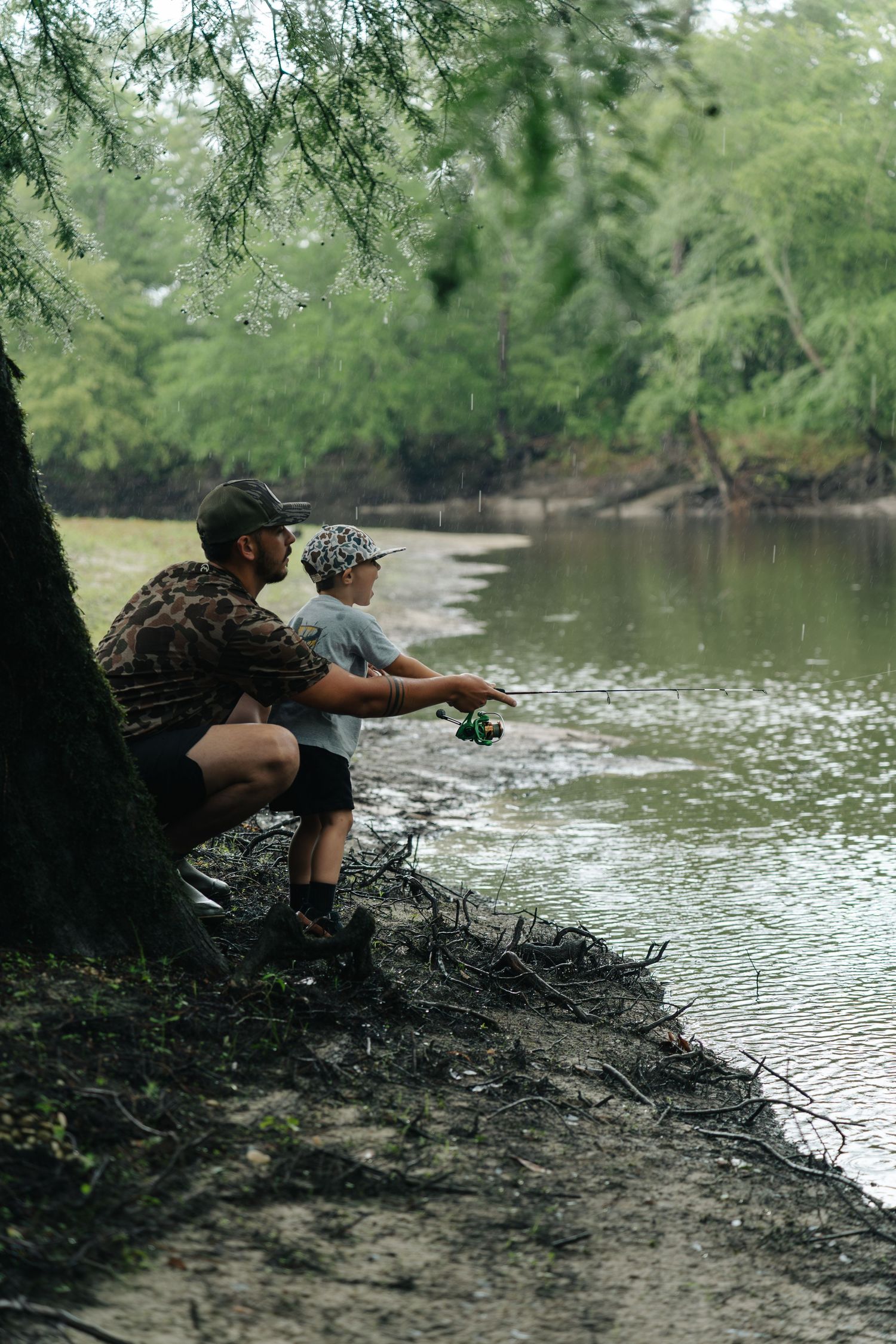 A father and young son fishing together on a tranquil river bank surrounded by lush green trees.