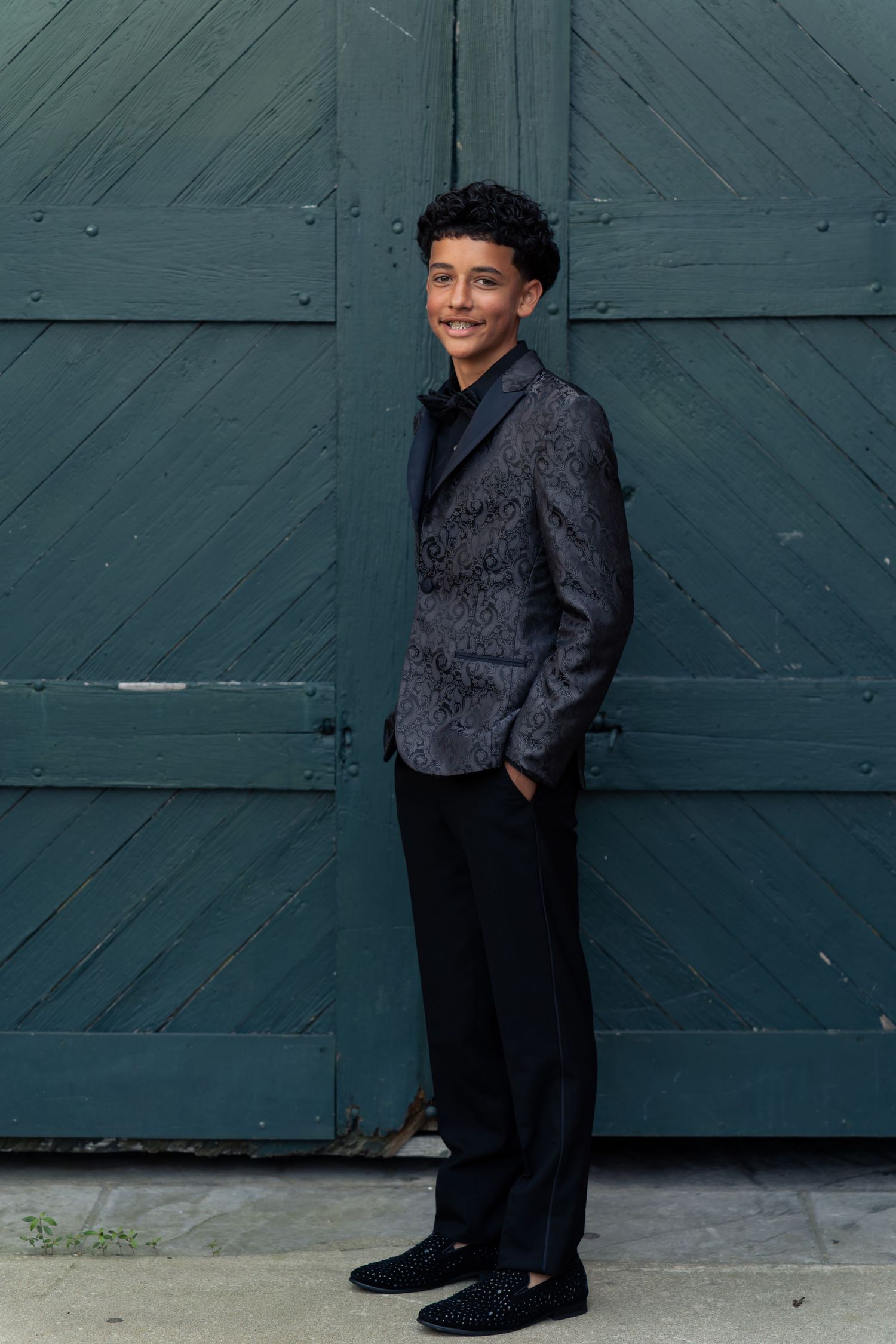 Young man in patterned blazer and black trousers smiling, standing before a teal wooden door.